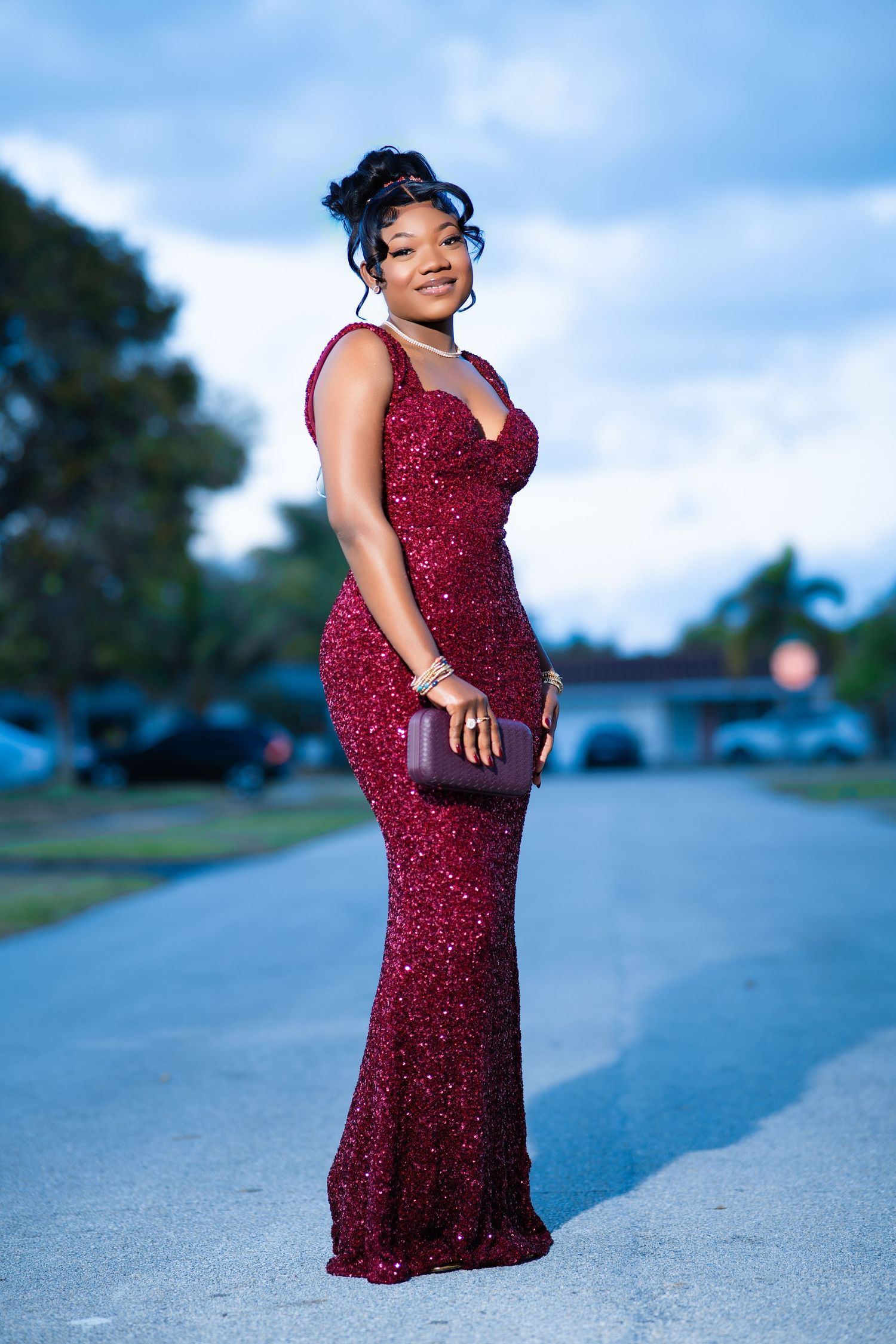 Young woman in a sparkling burgundy sequin gown holding a clutch purse, posing on a road at dusk.