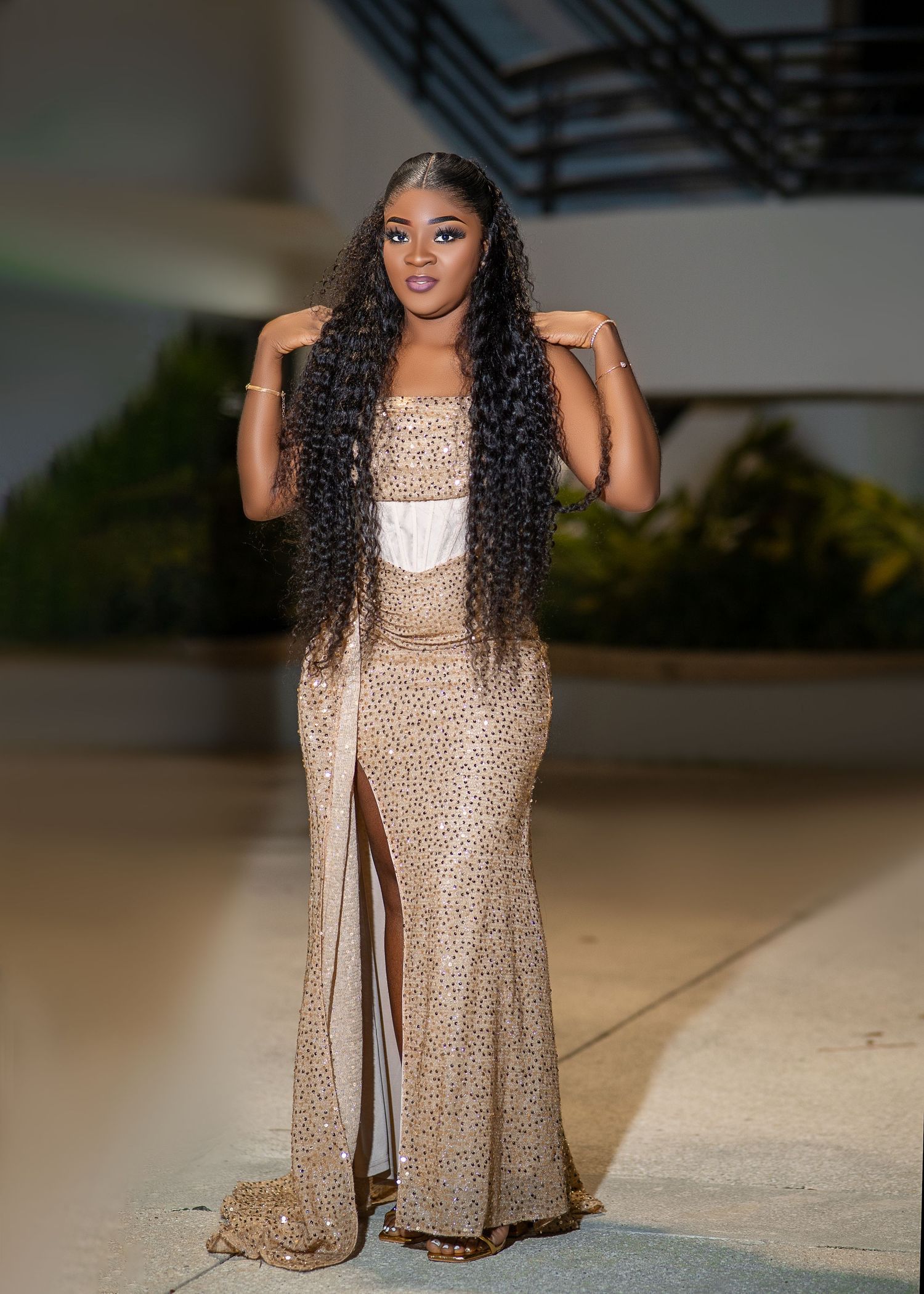 A person wearing a sparkly beige evening gown with a thigh-high slit and long wavy dark hair poses elegantly.