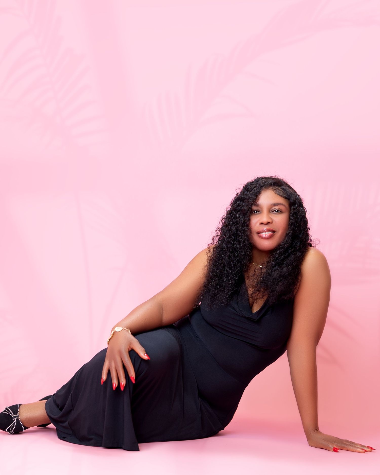 A woman in a black dress poses on a pink studio background with palm leaf shadows.