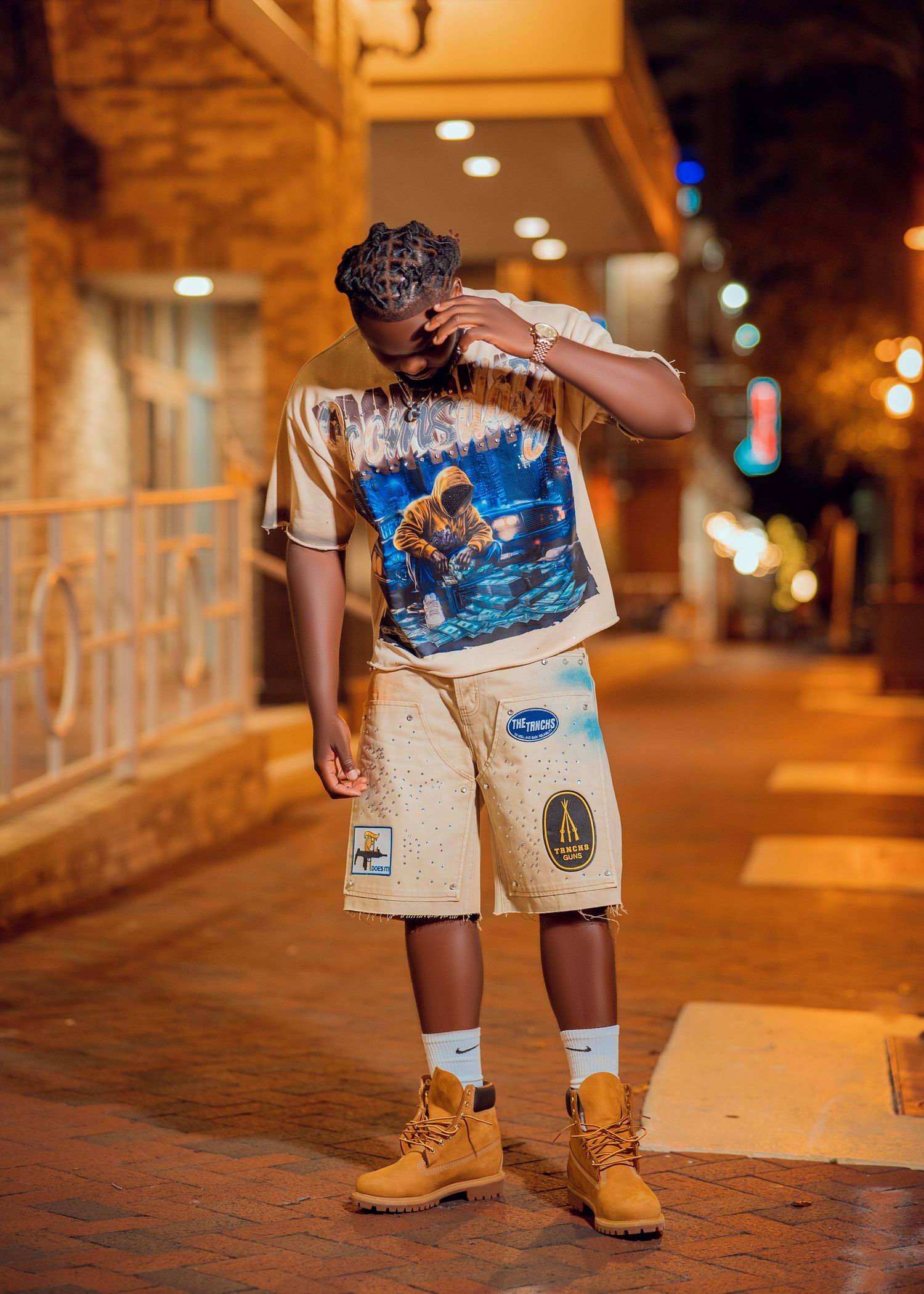 Young man posing on urban sidewalk at night wearing graphic tee, cargo shorts, white socks, and tan Timberland boots.