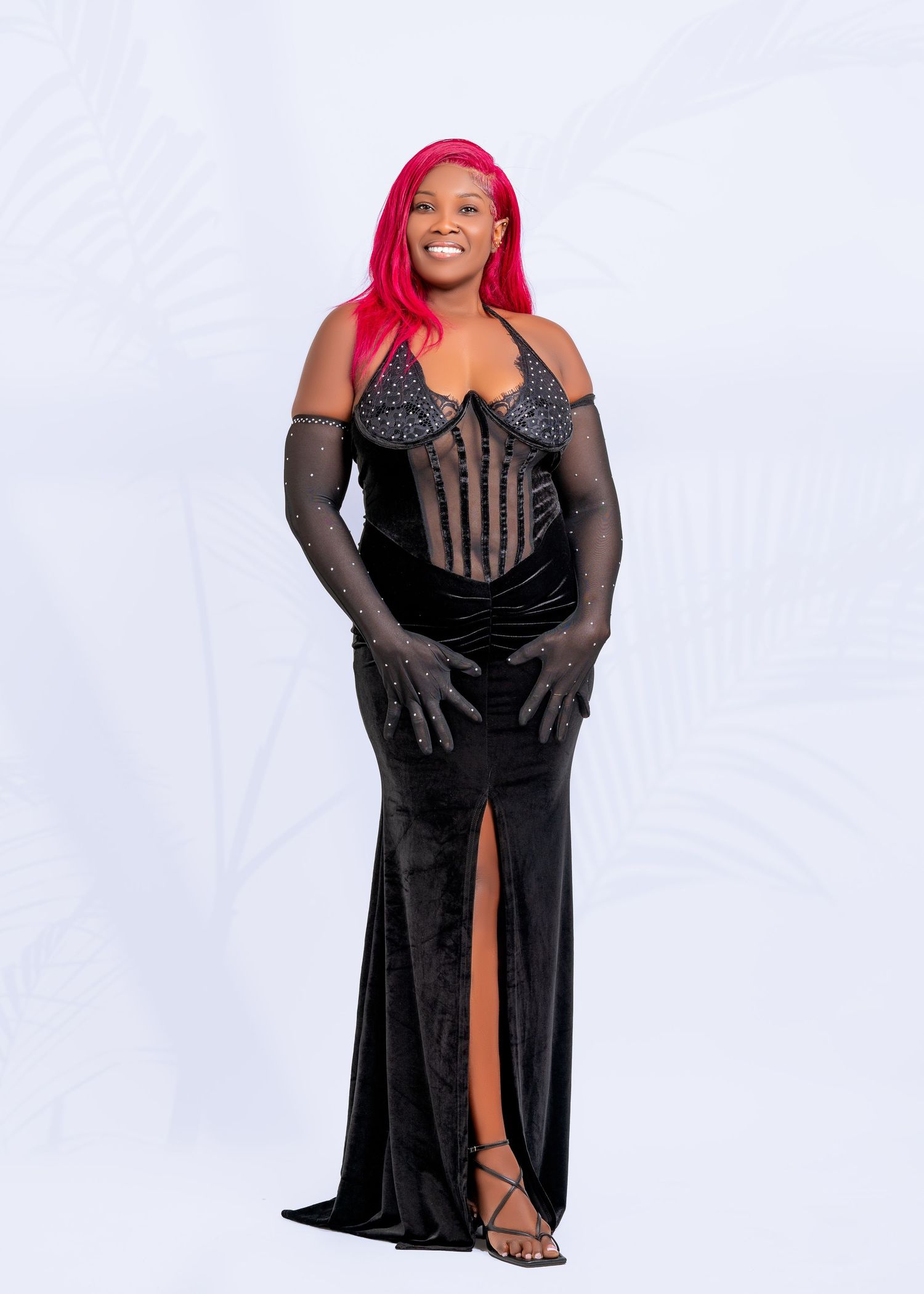 Woman in black evening gown with sheer details posing against white backdrop.