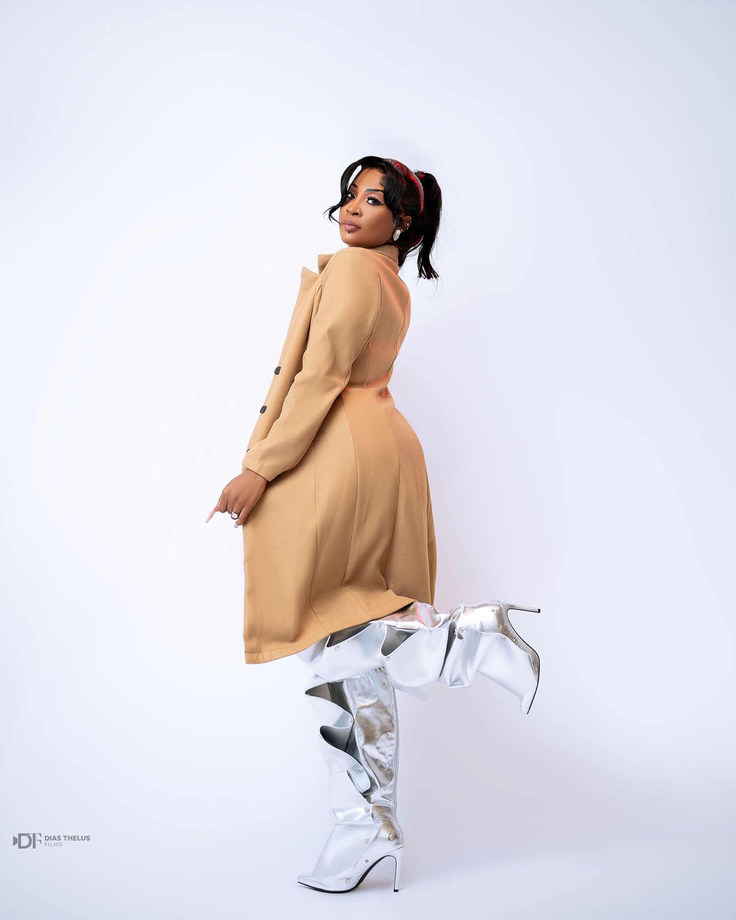 Woman in camel coat dress and silver knee-high boots poses gracefully against white background.