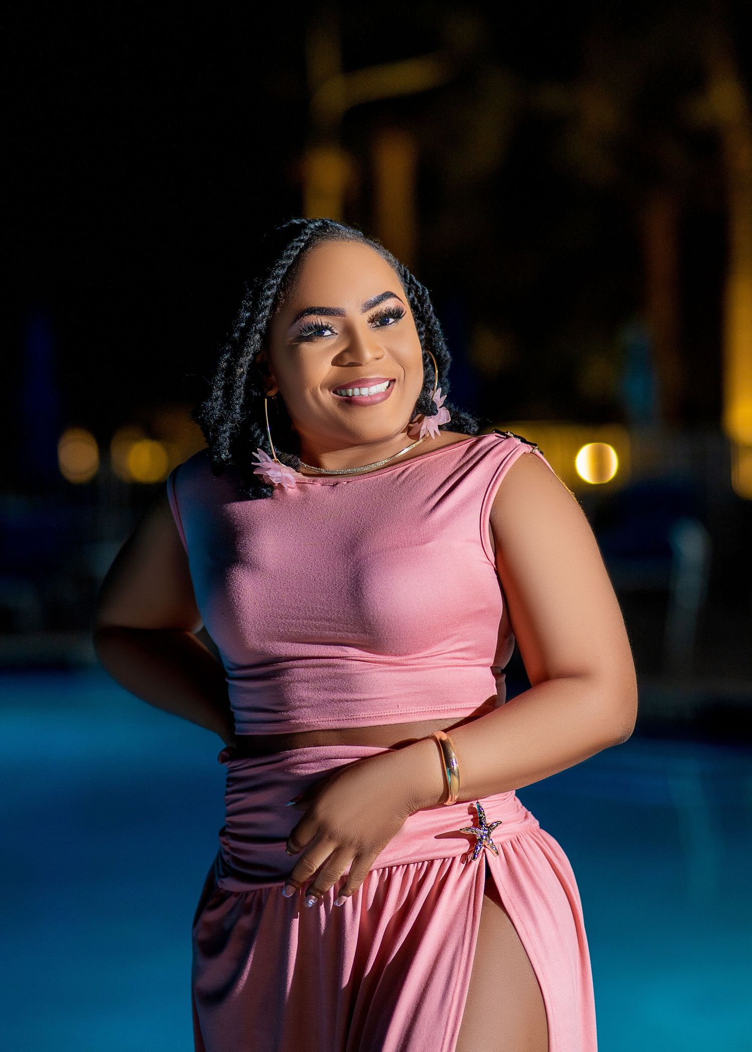 A person in a pink dress poses gracefully by a pool at night with blurred city lights in the background.