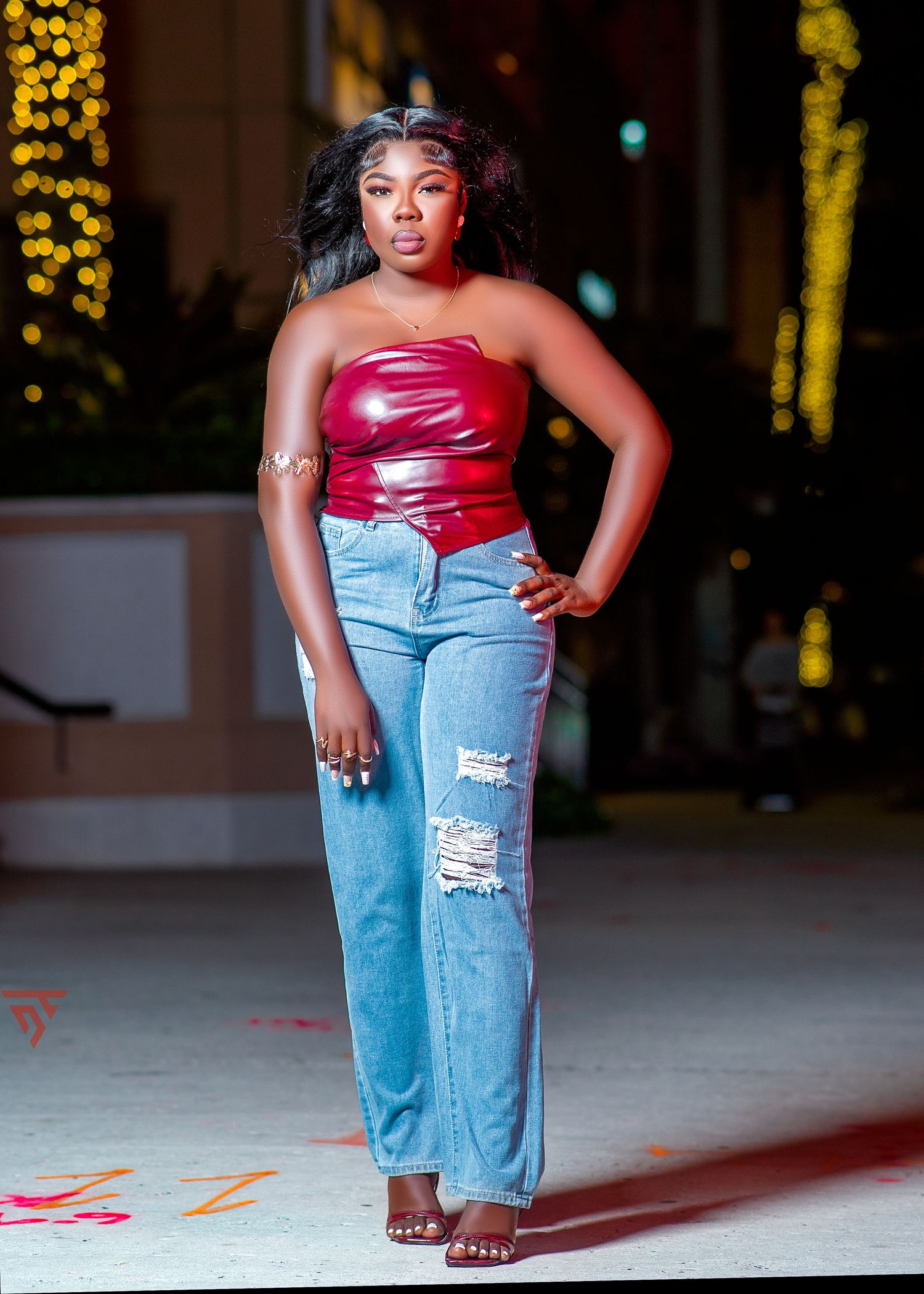 Fashion model poses in ripped light blue jeans and red top against urban nighttime backdrop with bokeh lights.