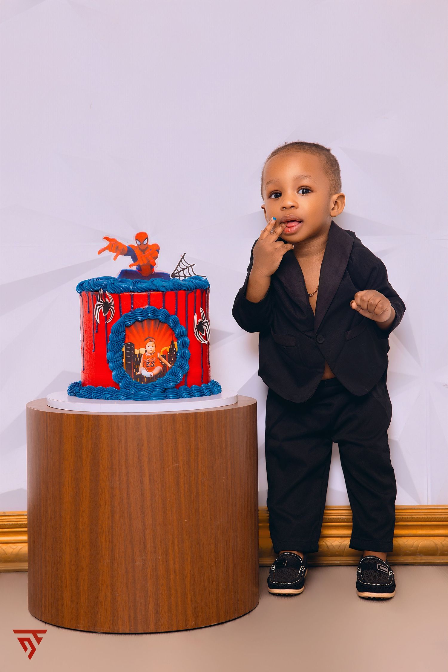 Baby in formal black suit sitting beside Spider-Man themed birthday cake.