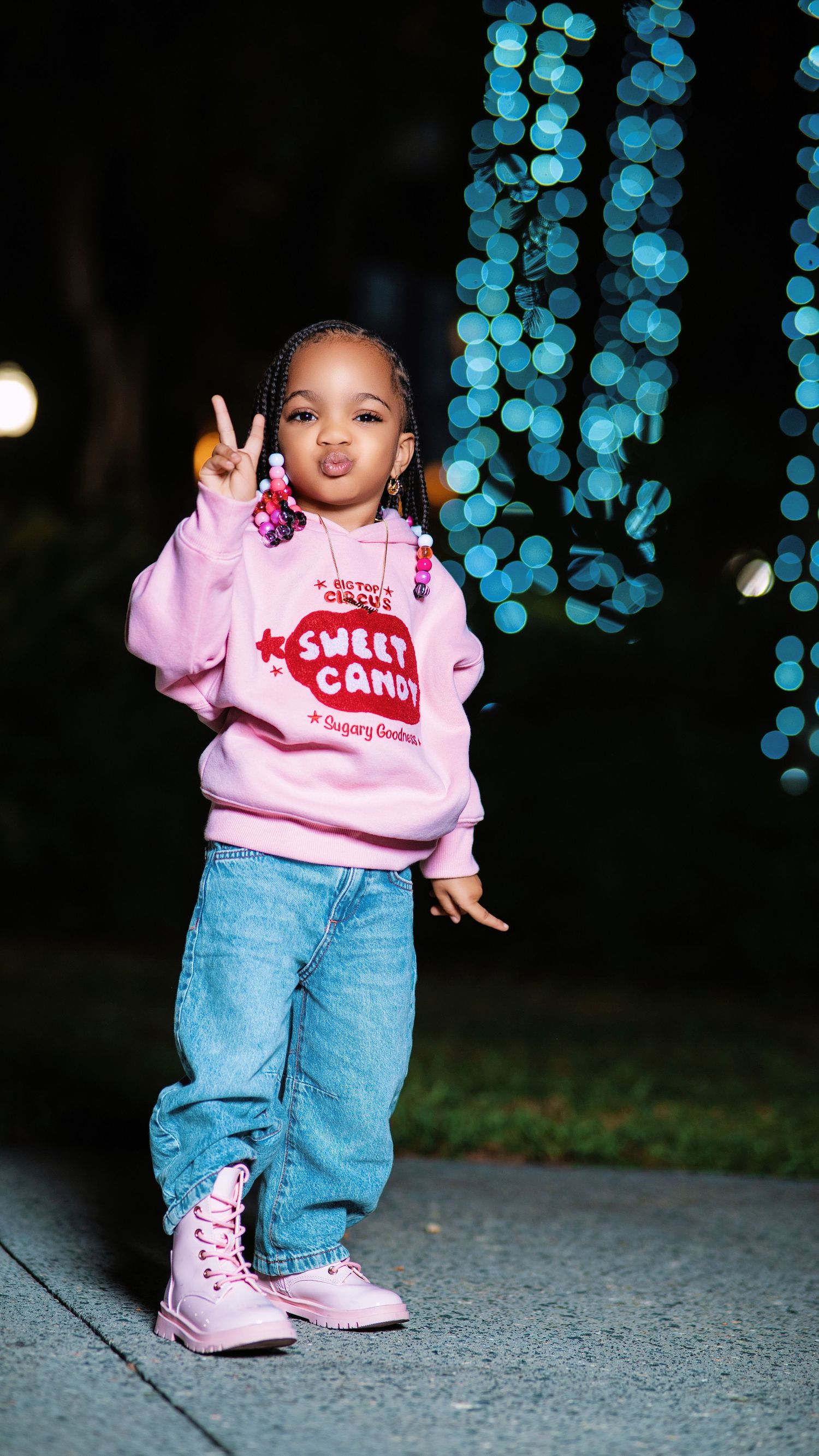 Child wearing pink Sweet Candy sweatshirt and jeans poses playfully outdoors at night.