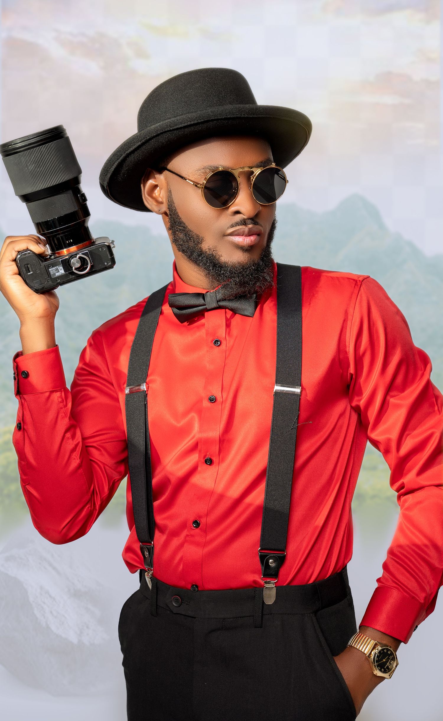 Photographer in red shirt, black suspenders and hat poses with vintage camera against scenic backdrop.