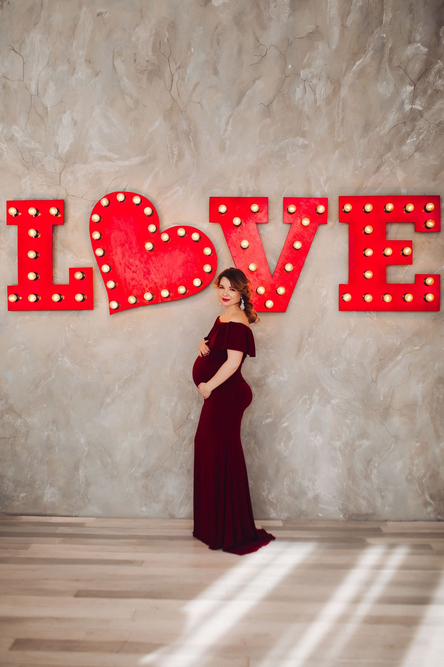 Maternity photo series with red light-up LOVE sign against textured wall.