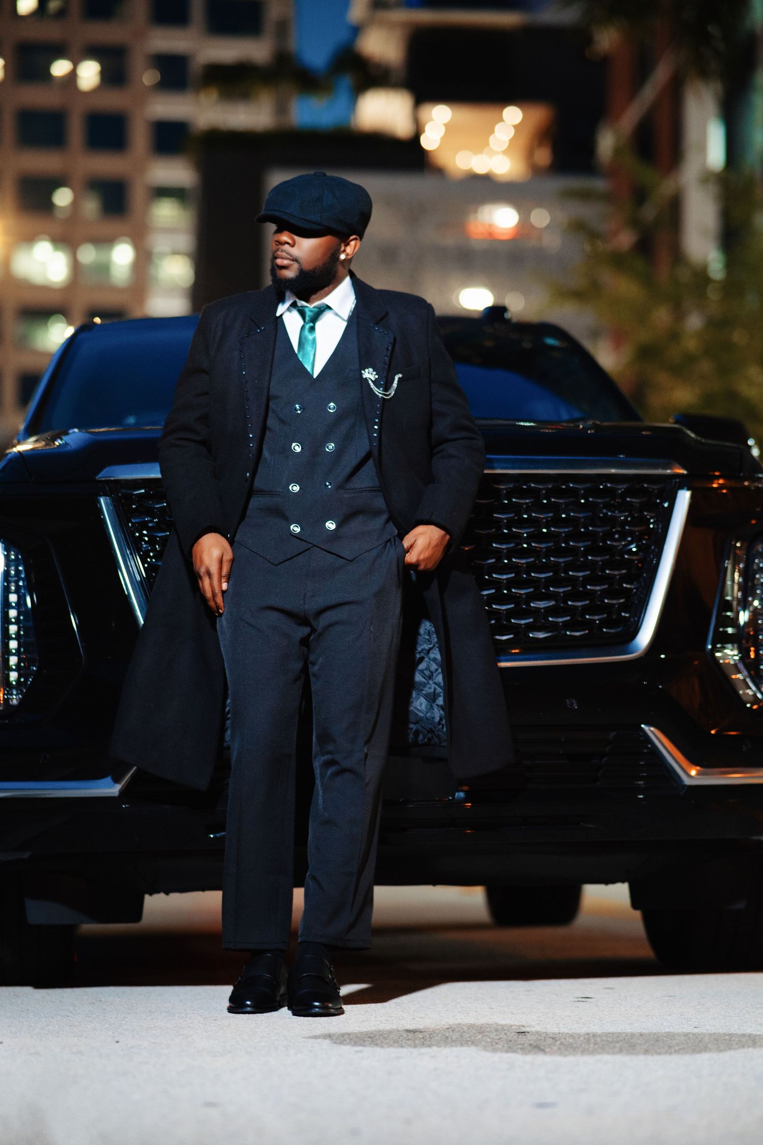 Elegant Black man in a tailored suit and flat cap poses confidently beside a black luxury SUV at night.