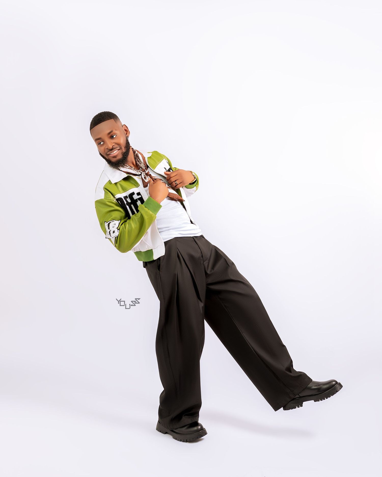 Young man posing confidently in green varsity jacket, wide-leg dark trousers, and black boots against white background.
