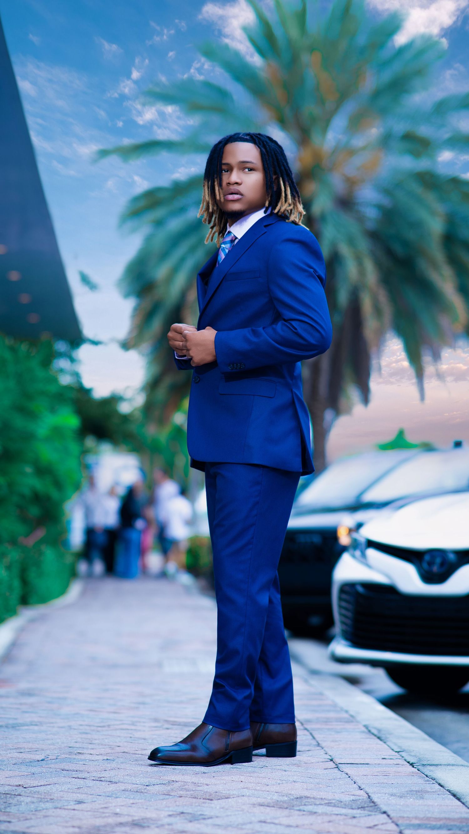 Young man in royal blue suit with dreadlocks poses confidently on brick path near palm trees and parked cars.
