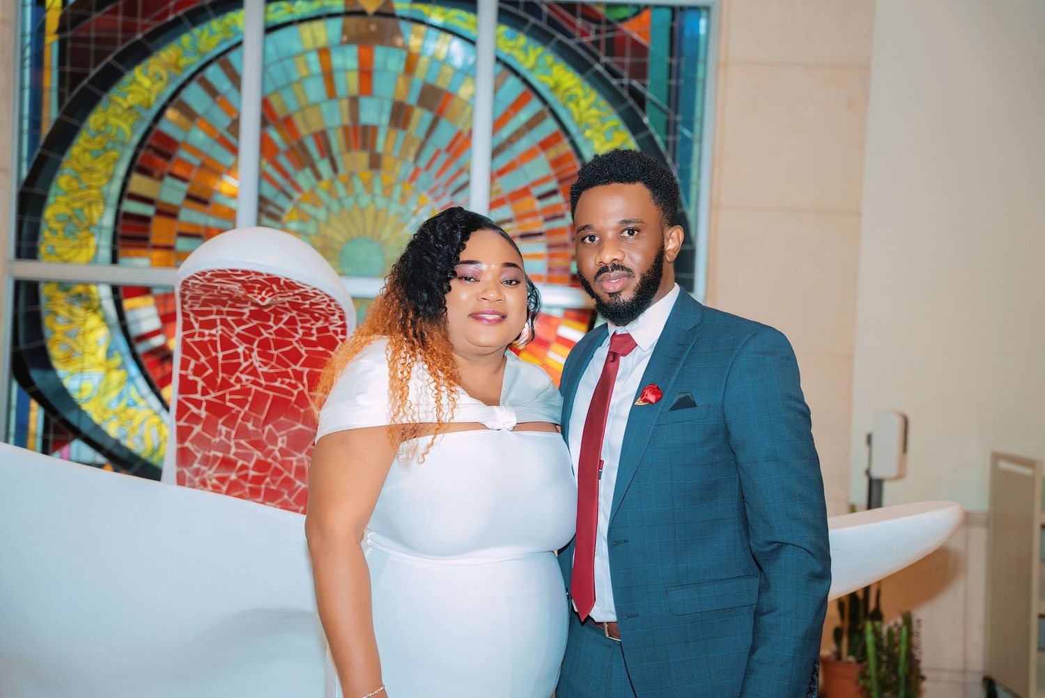 A couple poses in front of a vibrant stained glass wall design at an event.