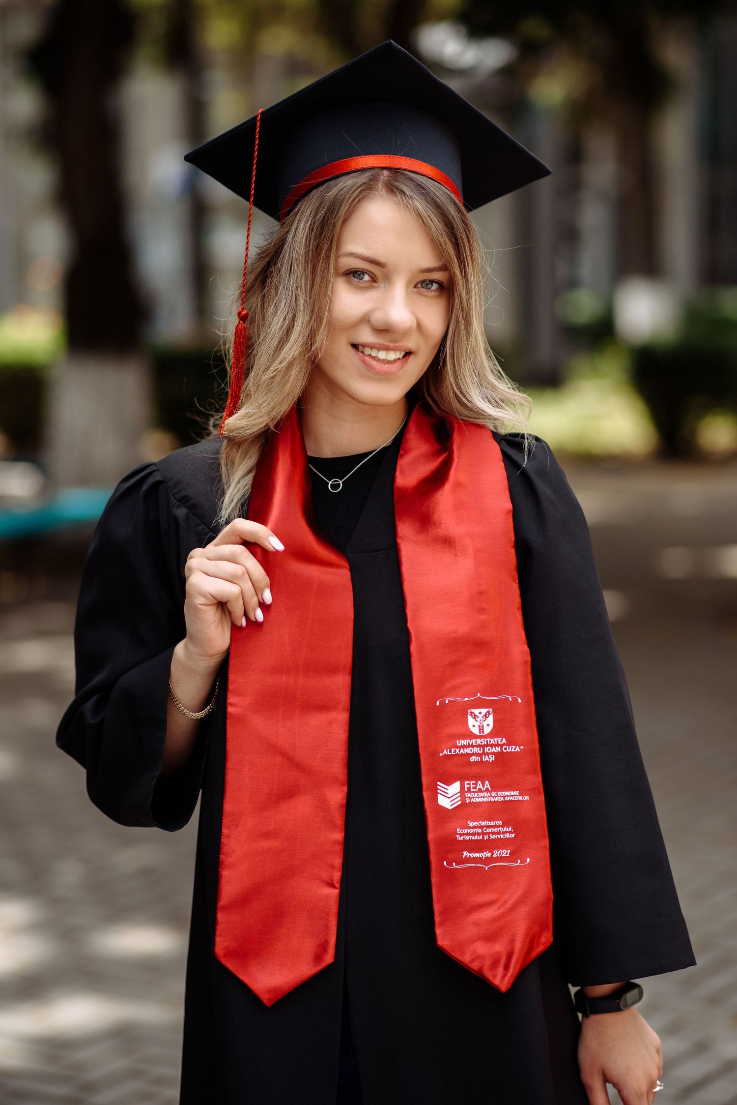 Graduate wearing black cap and gown with red stole smiles during photo session.