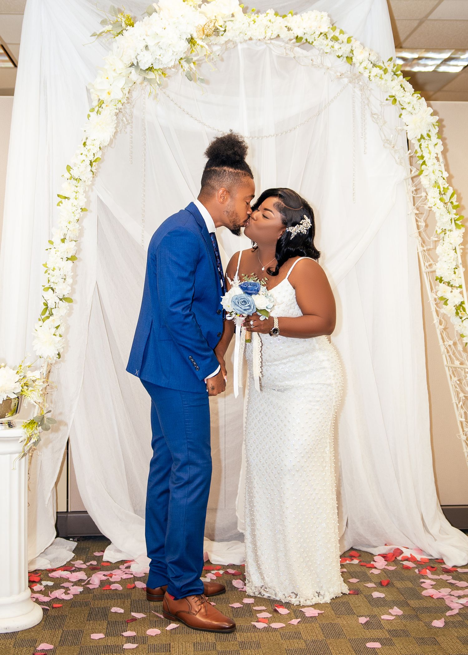 A romantic wedding scene with white floral arches and rose petals scattered on the floor.