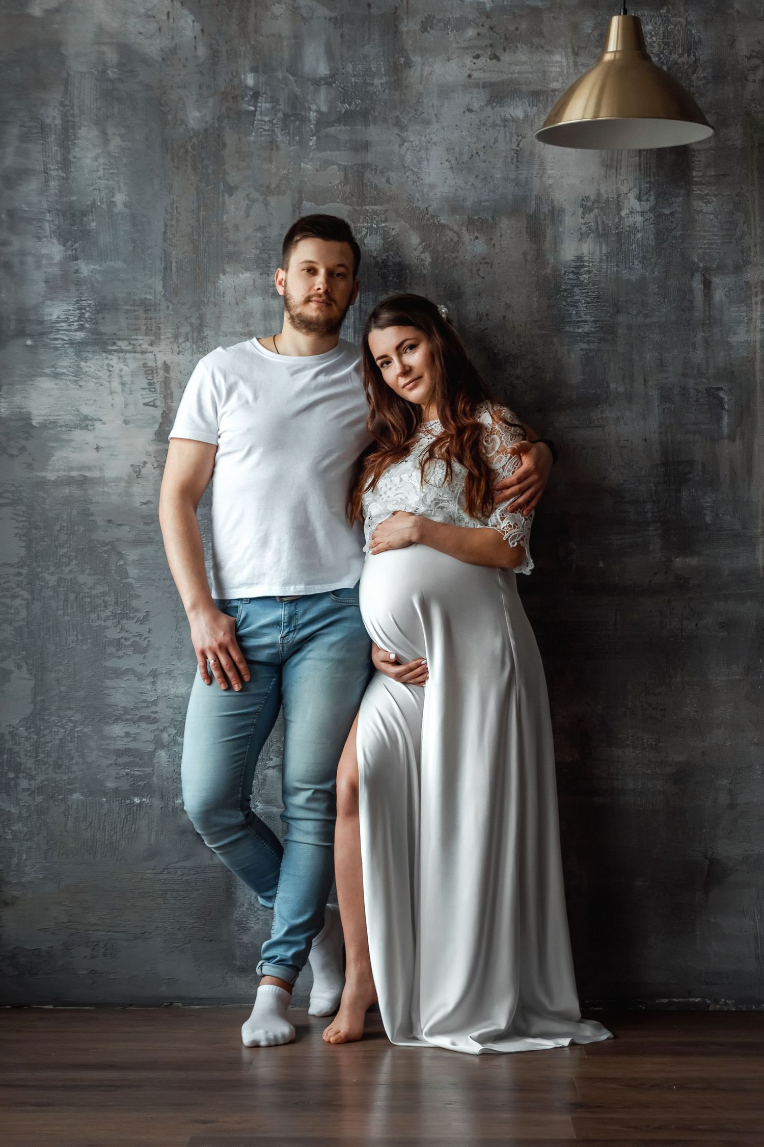 Romantic maternity photoshoot in white dress against grey wall with pendant lighting.