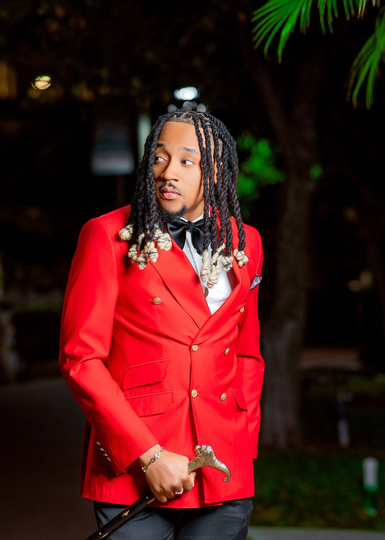 Person in bright red blazer with gold buttons poses confidently against dark background with palm leaves.
