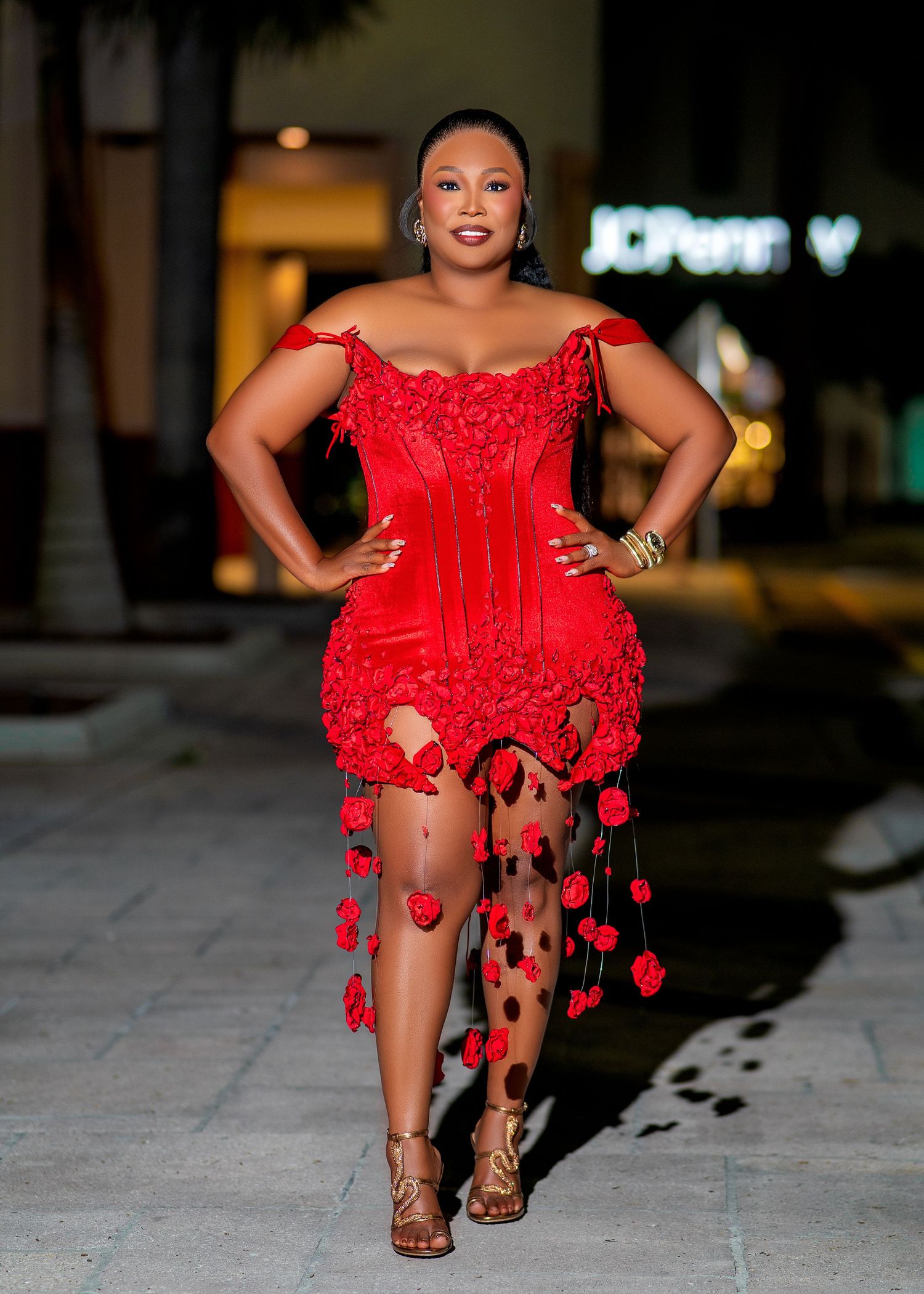 Woman in red floral corset mini dress with sheer rose-embellished train poses confidently outdoors at night.