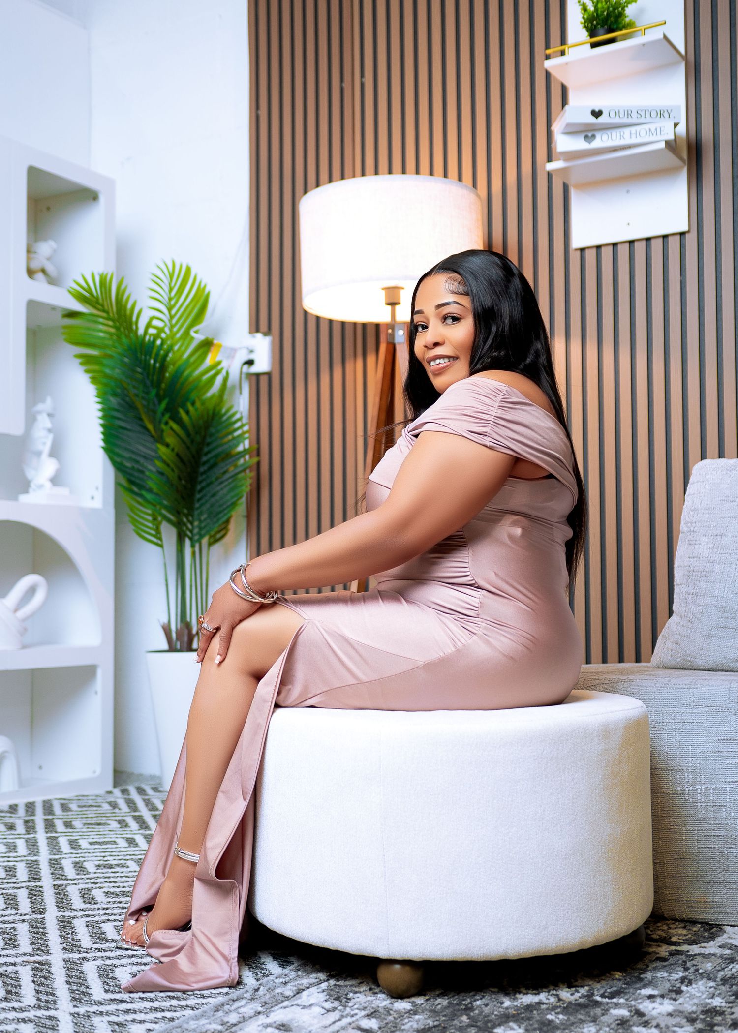 Woman in blush pink dress sitting on white ottoman in modern living room with wood panel wall and tropical plant.