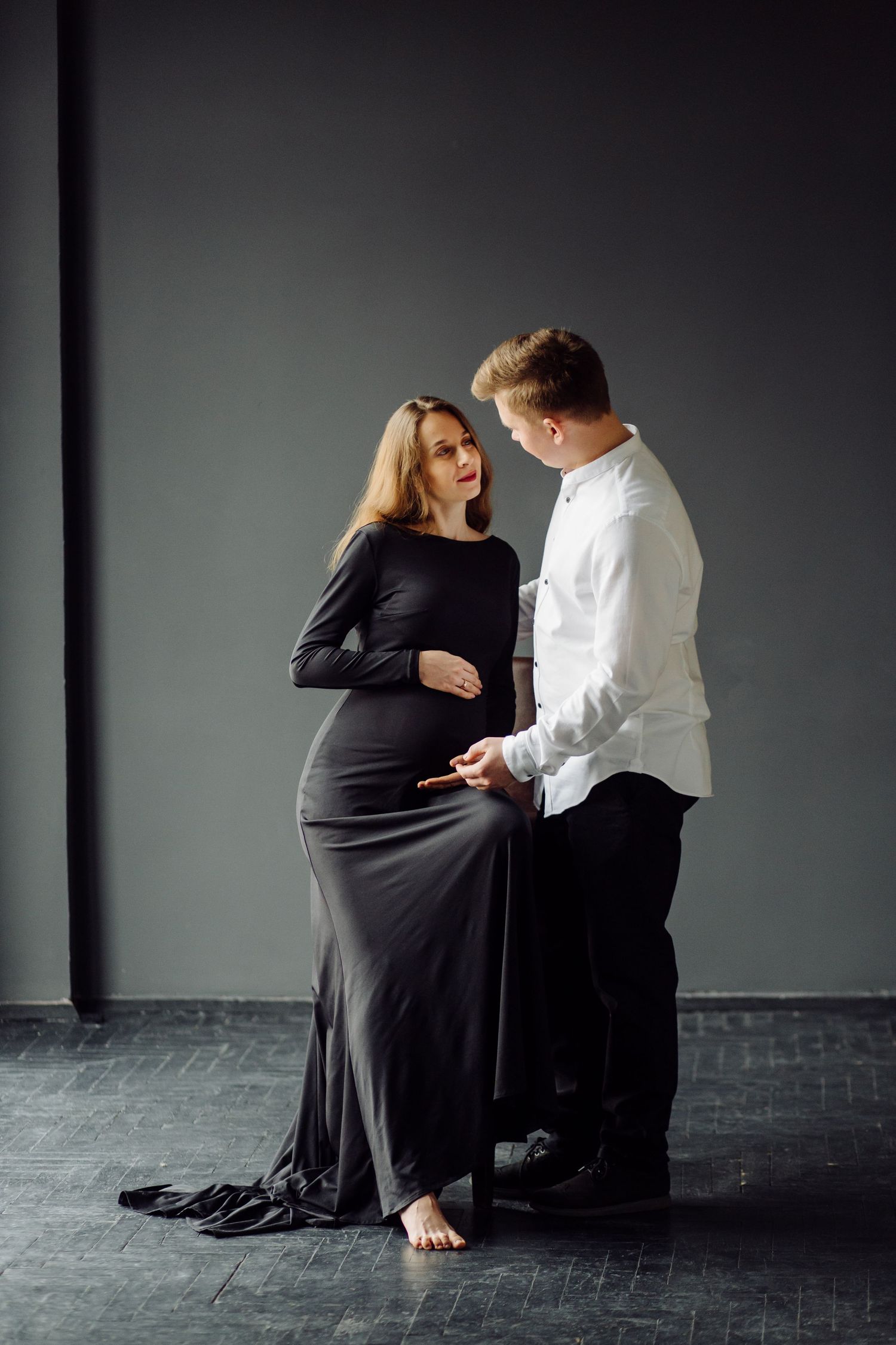Dramatic maternity photos in black dress against grey background showing tender moments.