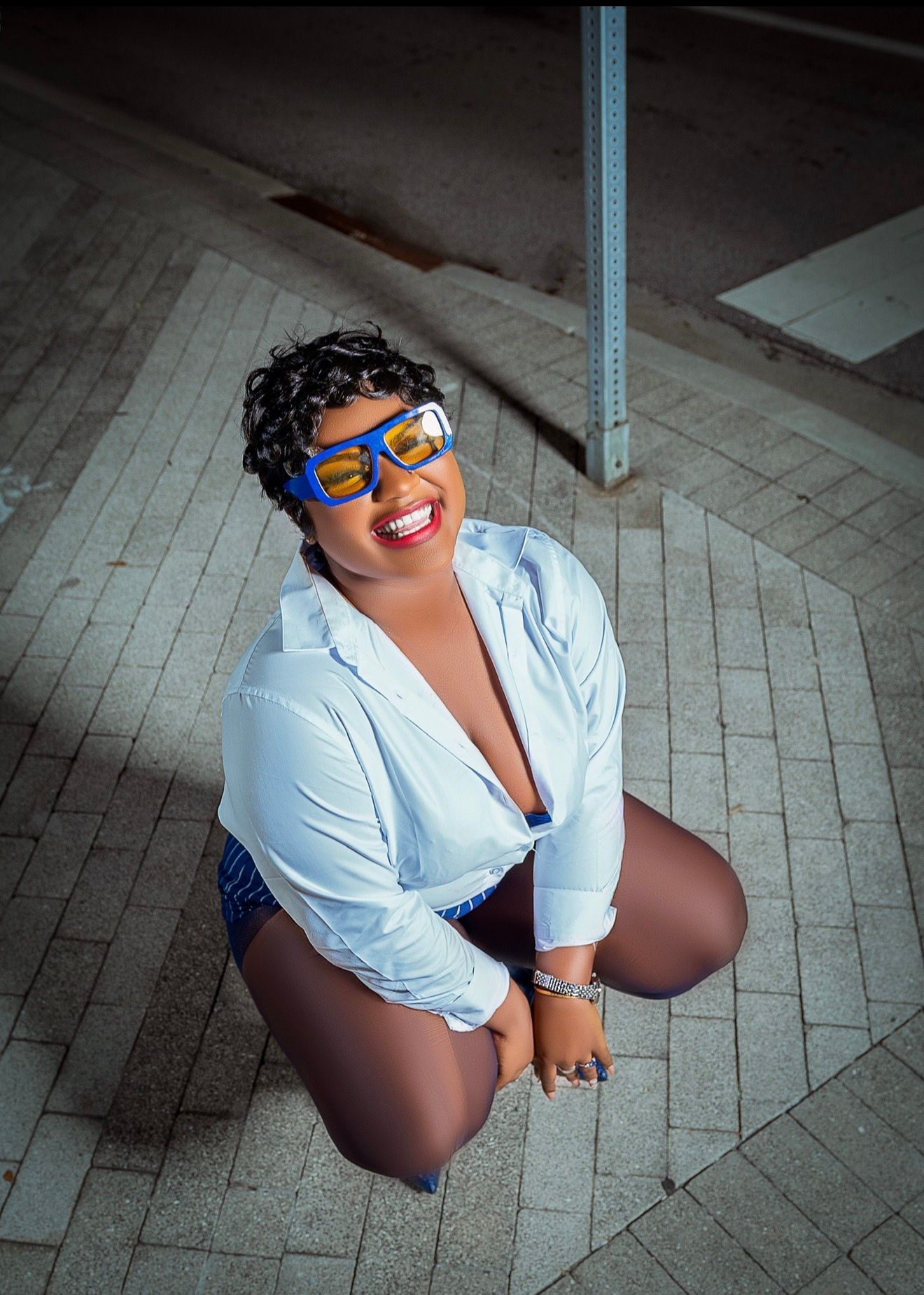 Smiling woman in white shirt and blue glasses posing on sidewalk, looking up at camera with confidence.