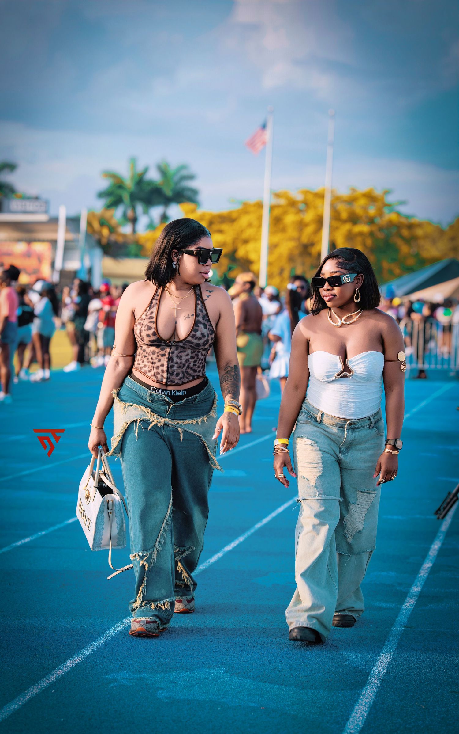 Two friends walking on a blue track at an outdoor event with palm trees and flags in the background.