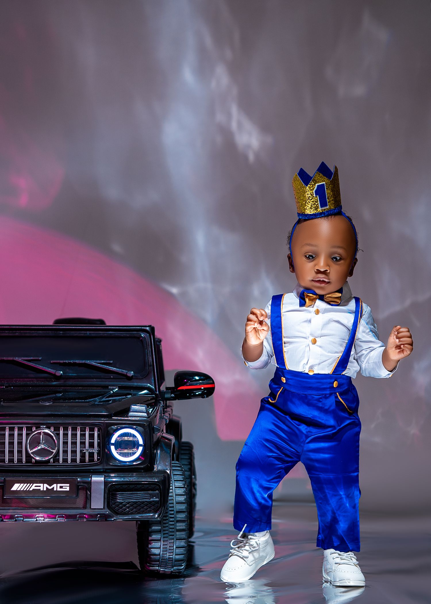 Adorable toddler boy in royal blue suspenders, bow tie, and gold crown poses beside a toy Mercedes-AMG G-Wagon.