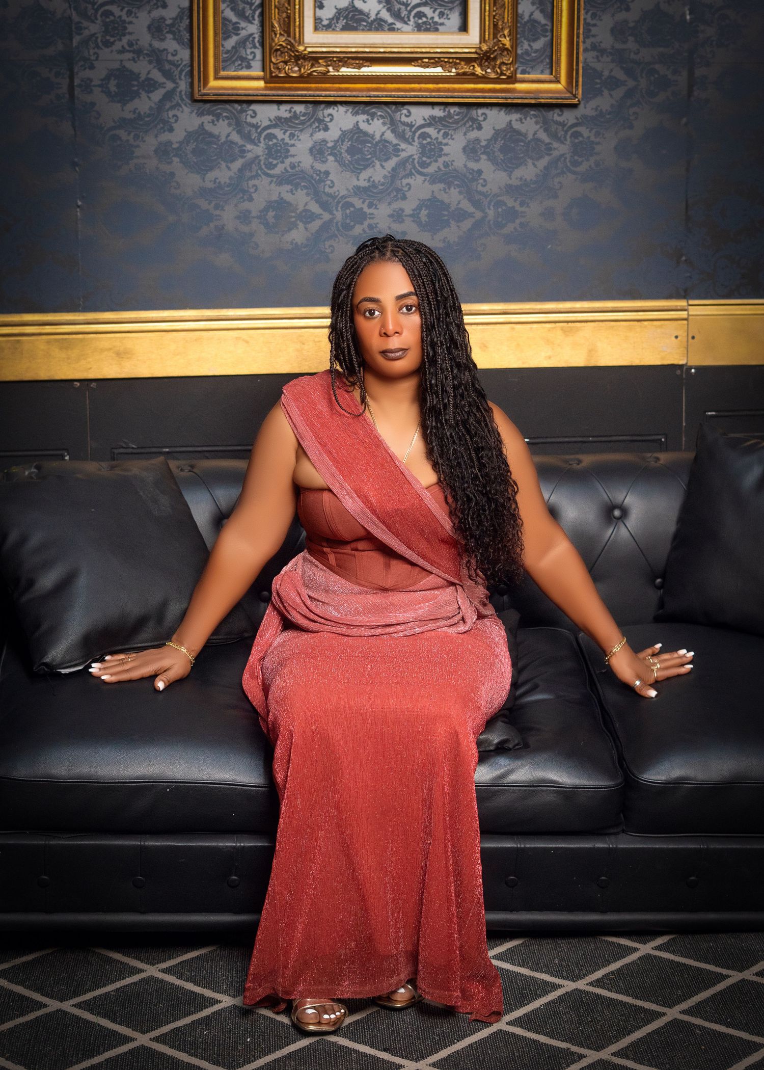 Woman in red velvet dress poses on black leather couch in elegant indoor setting.
