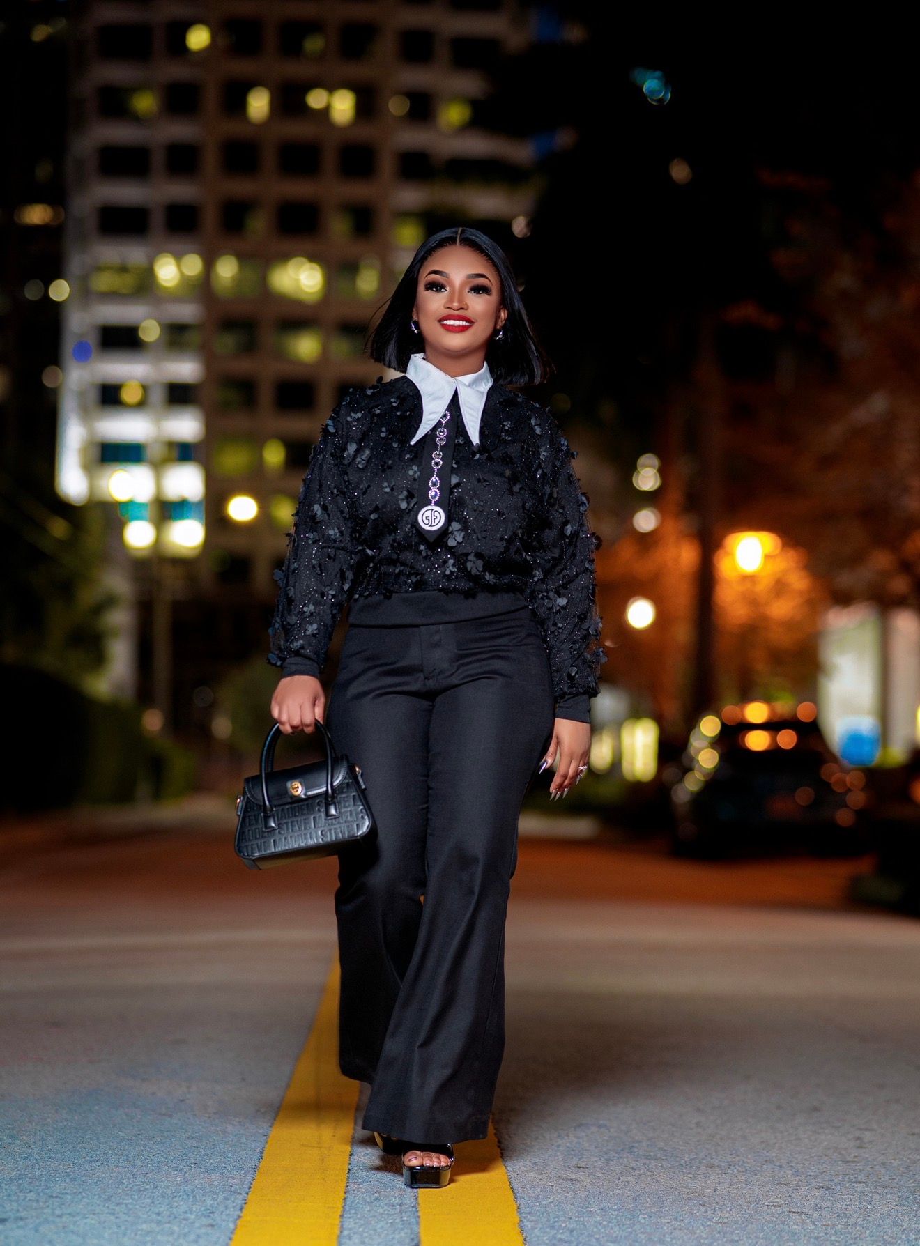 Woman in black lace top, wide-leg pants, and handbag walking confidently on a city street at night.