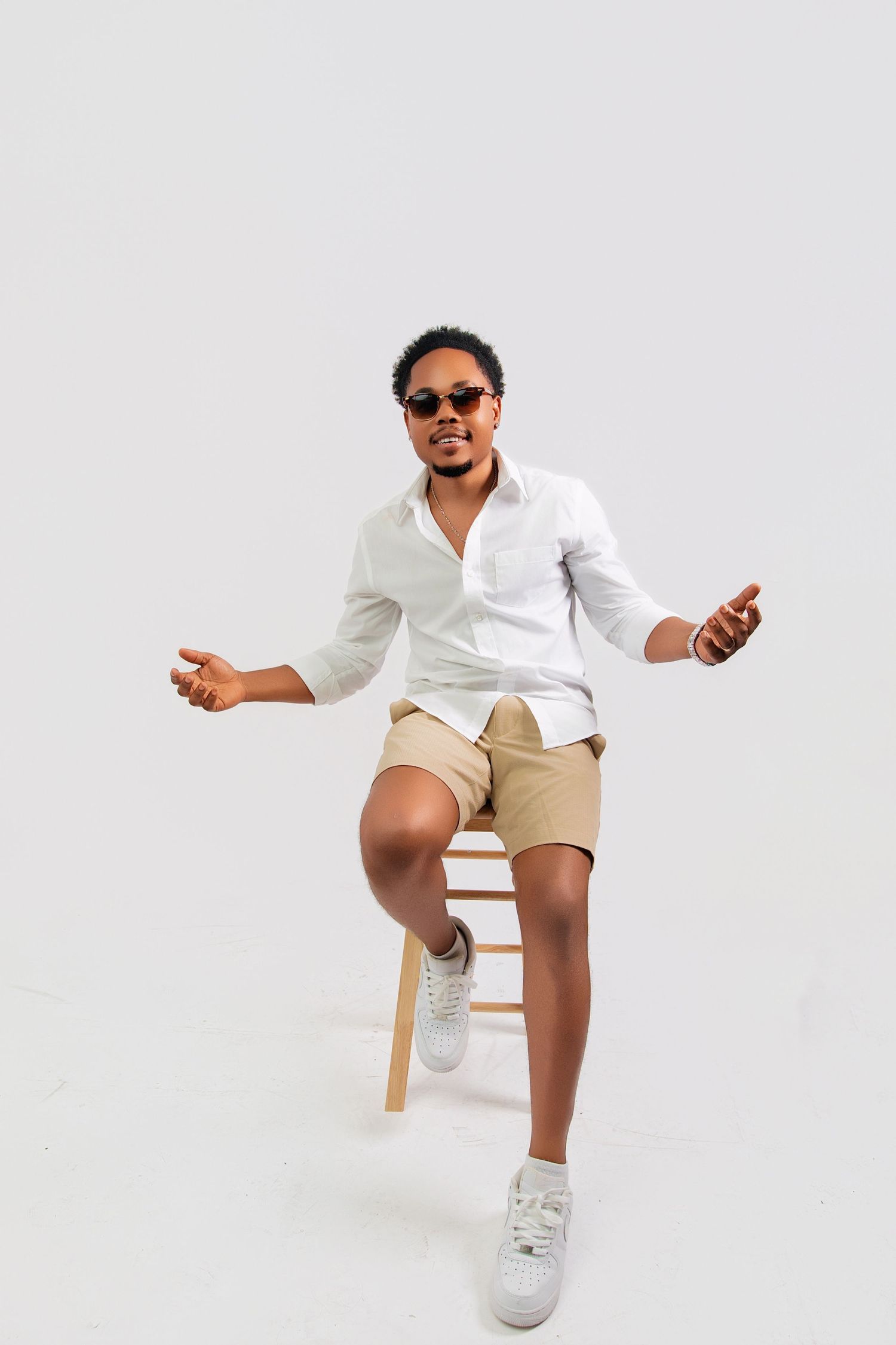 Stylish Black man sitting on wooden stool wearing white shirt, khaki shorts, sunglasses, and white sneakers on white background.