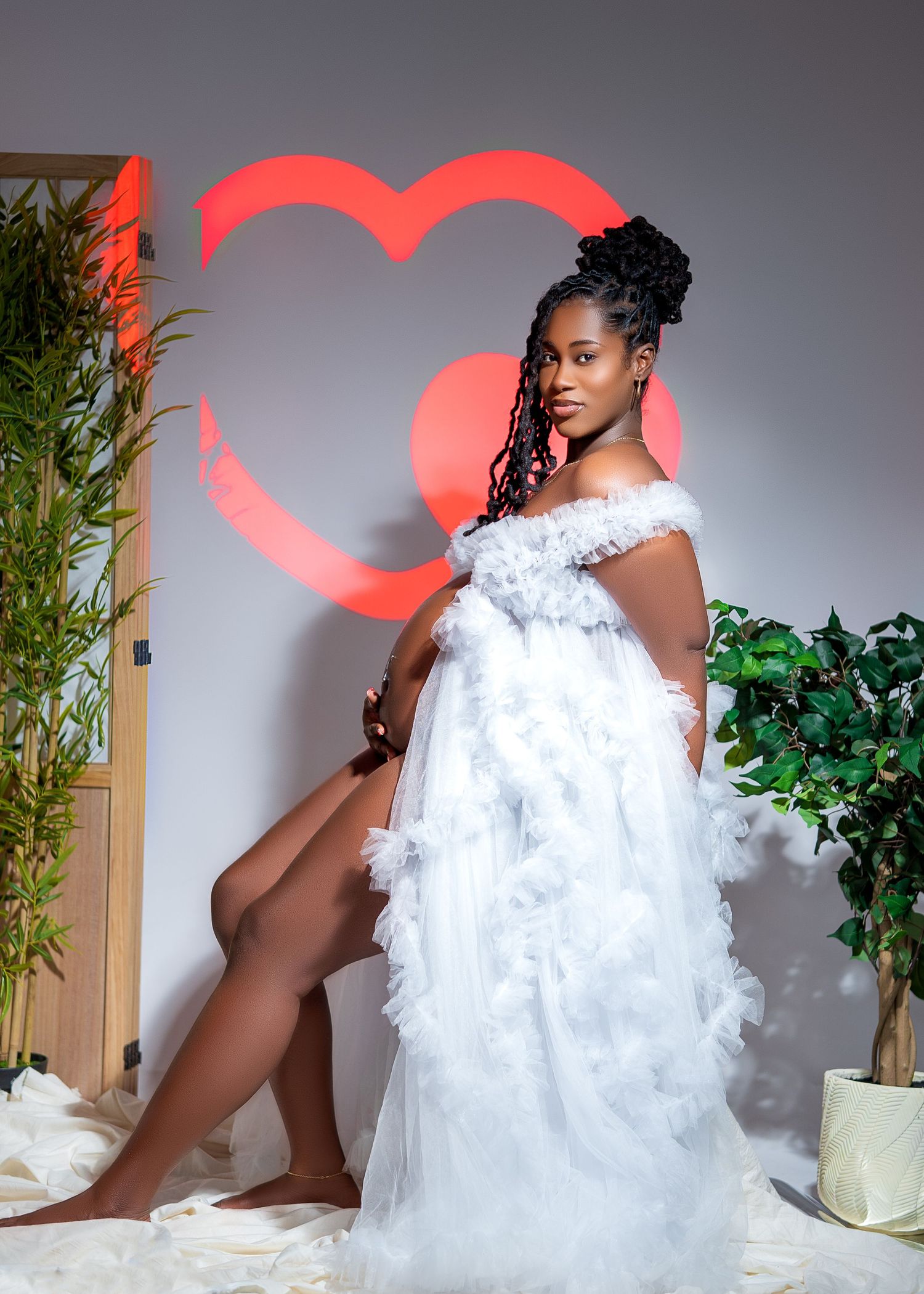 Pregnant woman in white ruffled off-shoulder gown posing elegantly in front of a red heart backdrop.