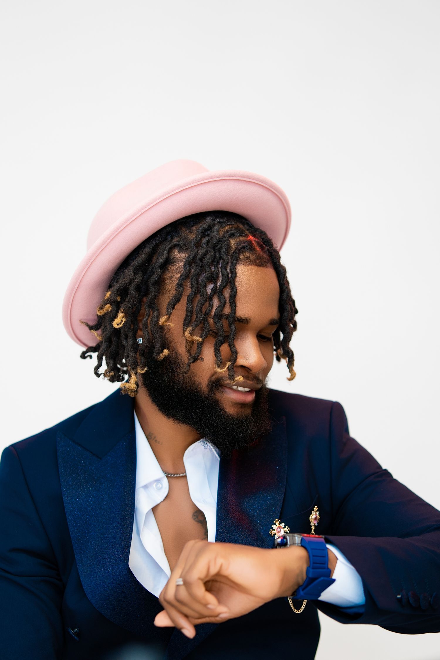 Stylish man with dreadlocks wearing a pink hat and navy suit checks his watch against a white background.