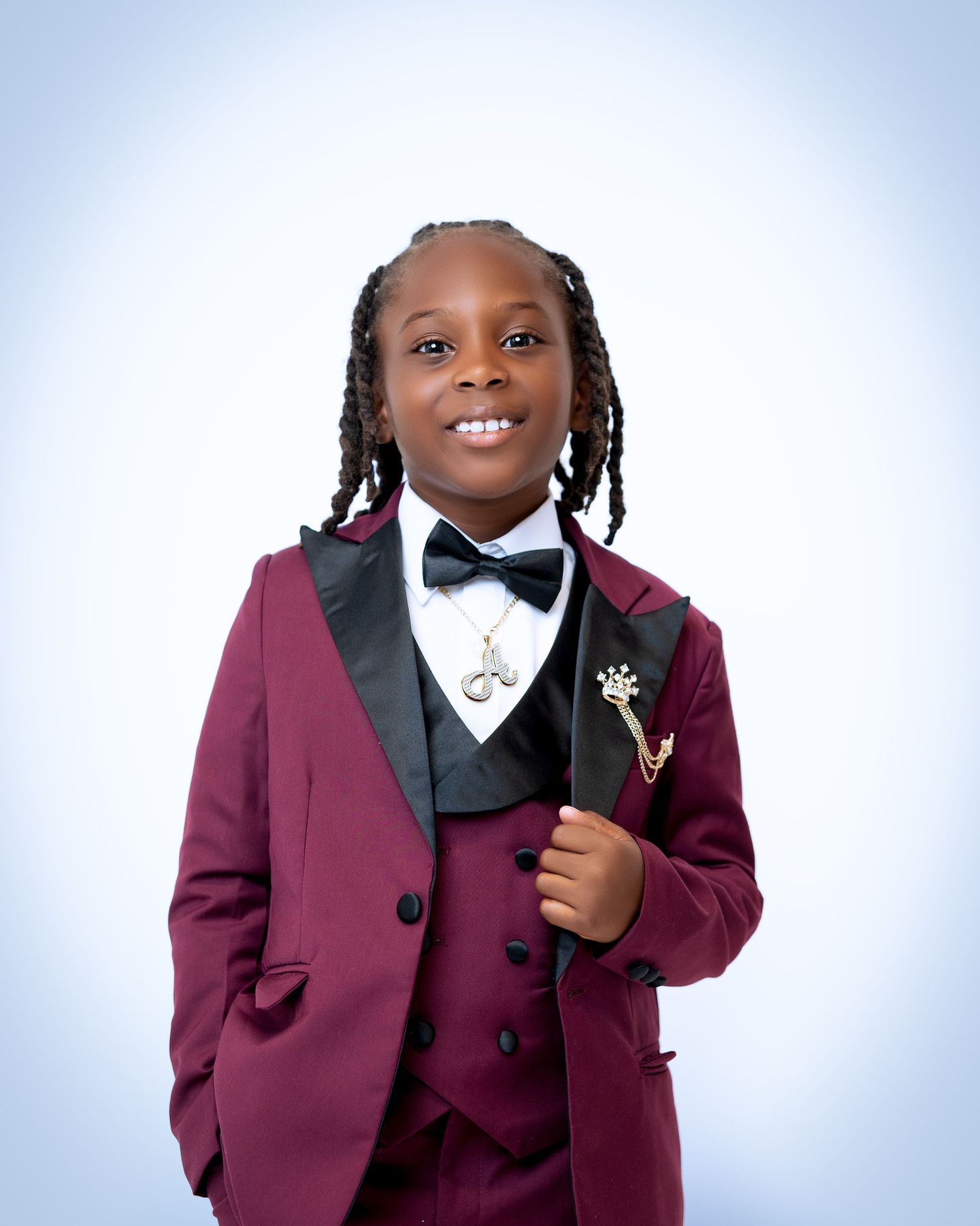 Young boy in burgundy tuxedo with black bow tie and braids, holding a cane, smiling for a formal portrait.