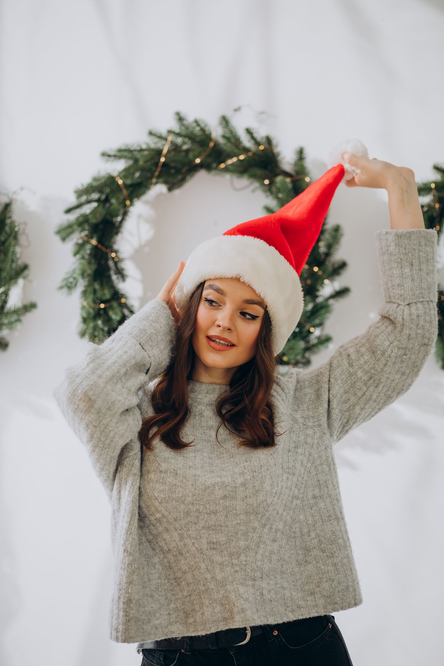 Person in gray sweater and Santa hat posing playfully against white background with holiday wreath decor.