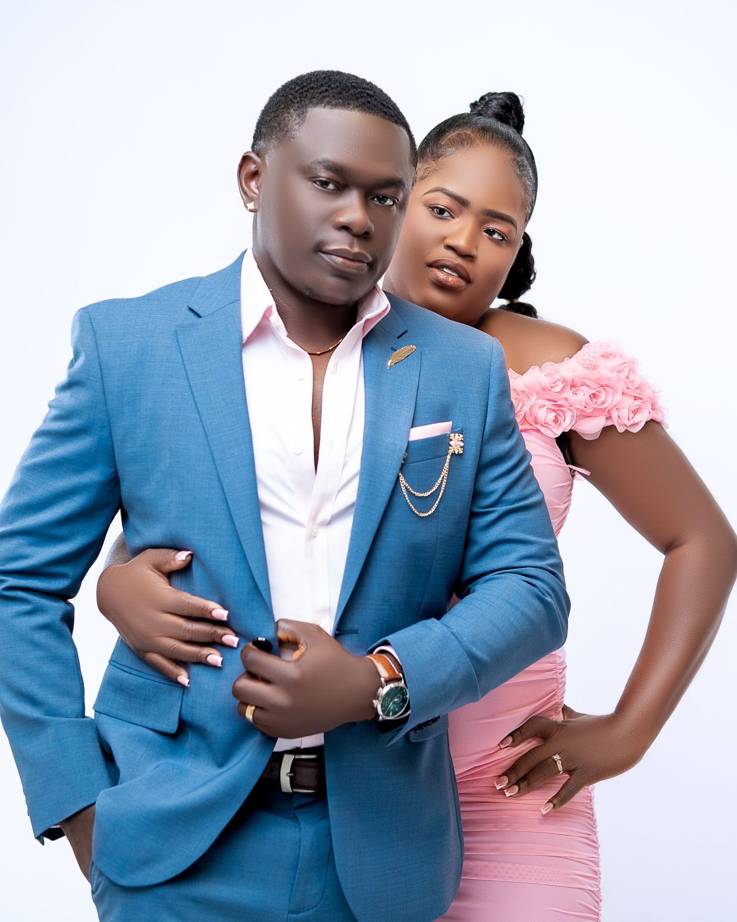 A young couple poses elegantly, man in blue suit and woman in pink dress, against a white background.