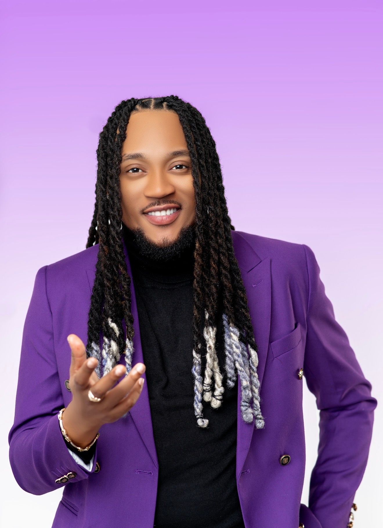 Smiling man with long braided locs wearing a purple blazer over black shirt poses against a purple gradient background.