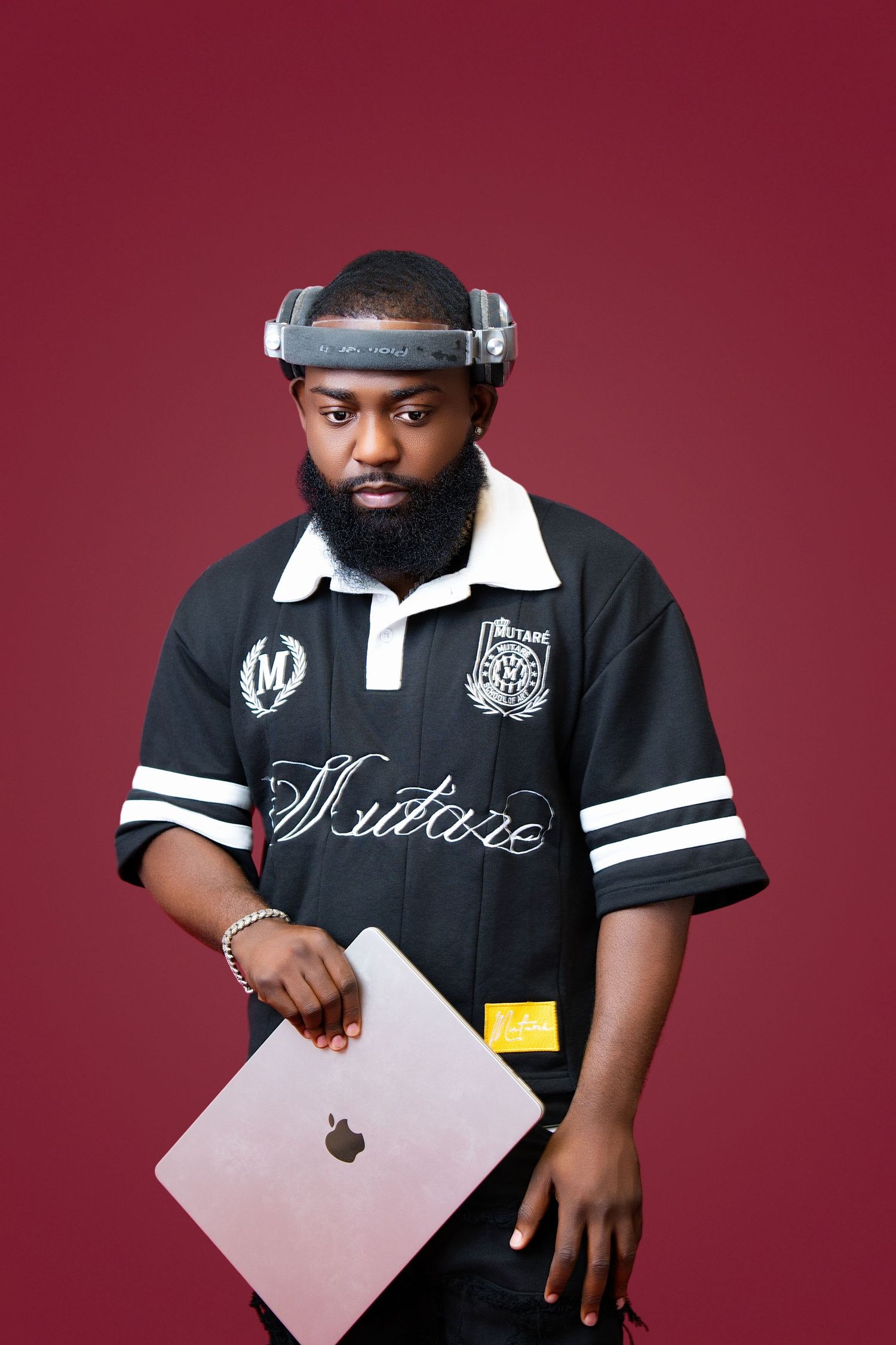 Man with beard wearing black jersey holds MacBook, futuristic headset on head, posing against dark red background.