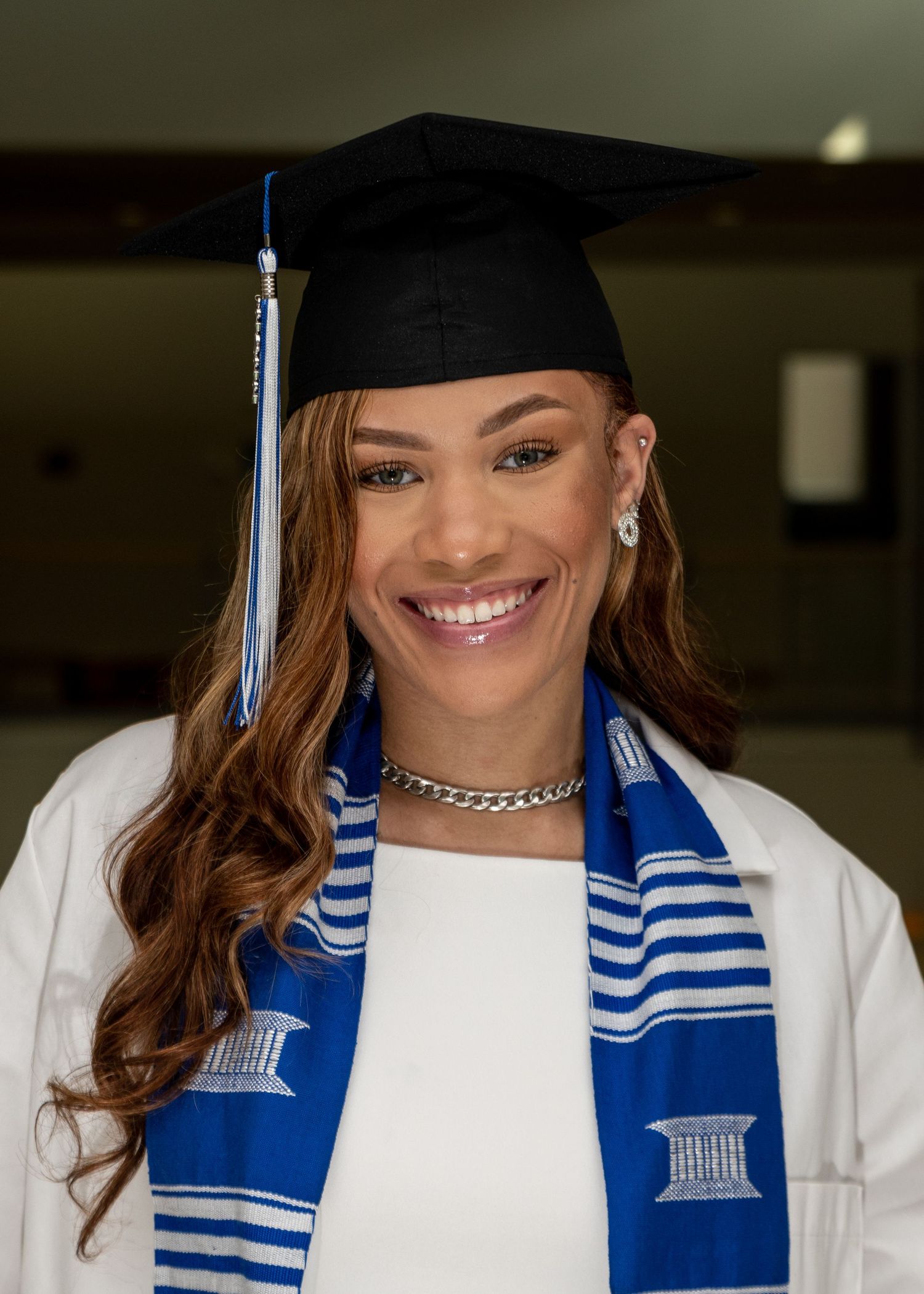 Graduate in white gown and blue striped stole poses indoors for commencement photos.