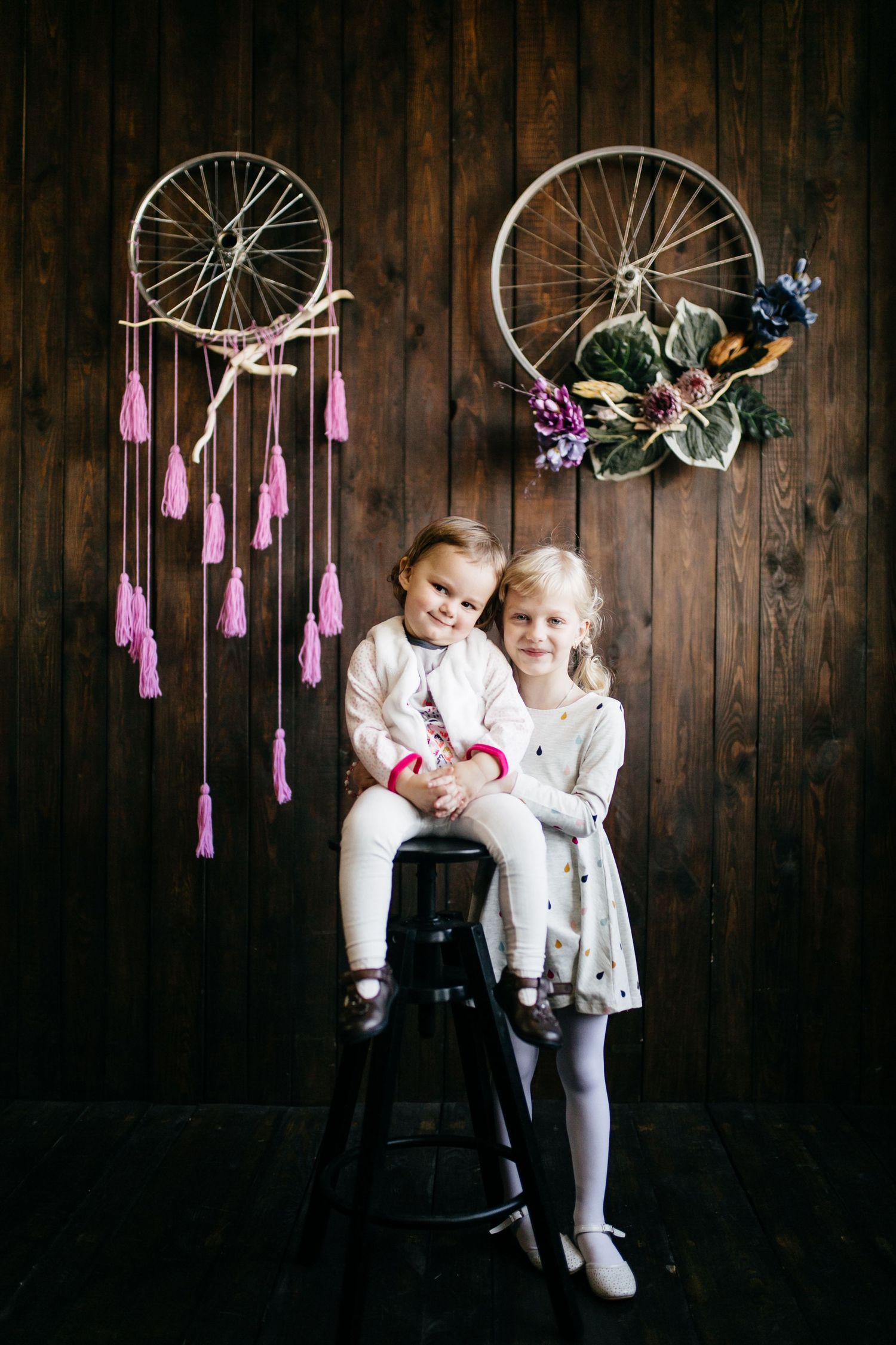 Two children in white outfits pose under hanging pink decorations and vintage bicycles on dark wood wall.