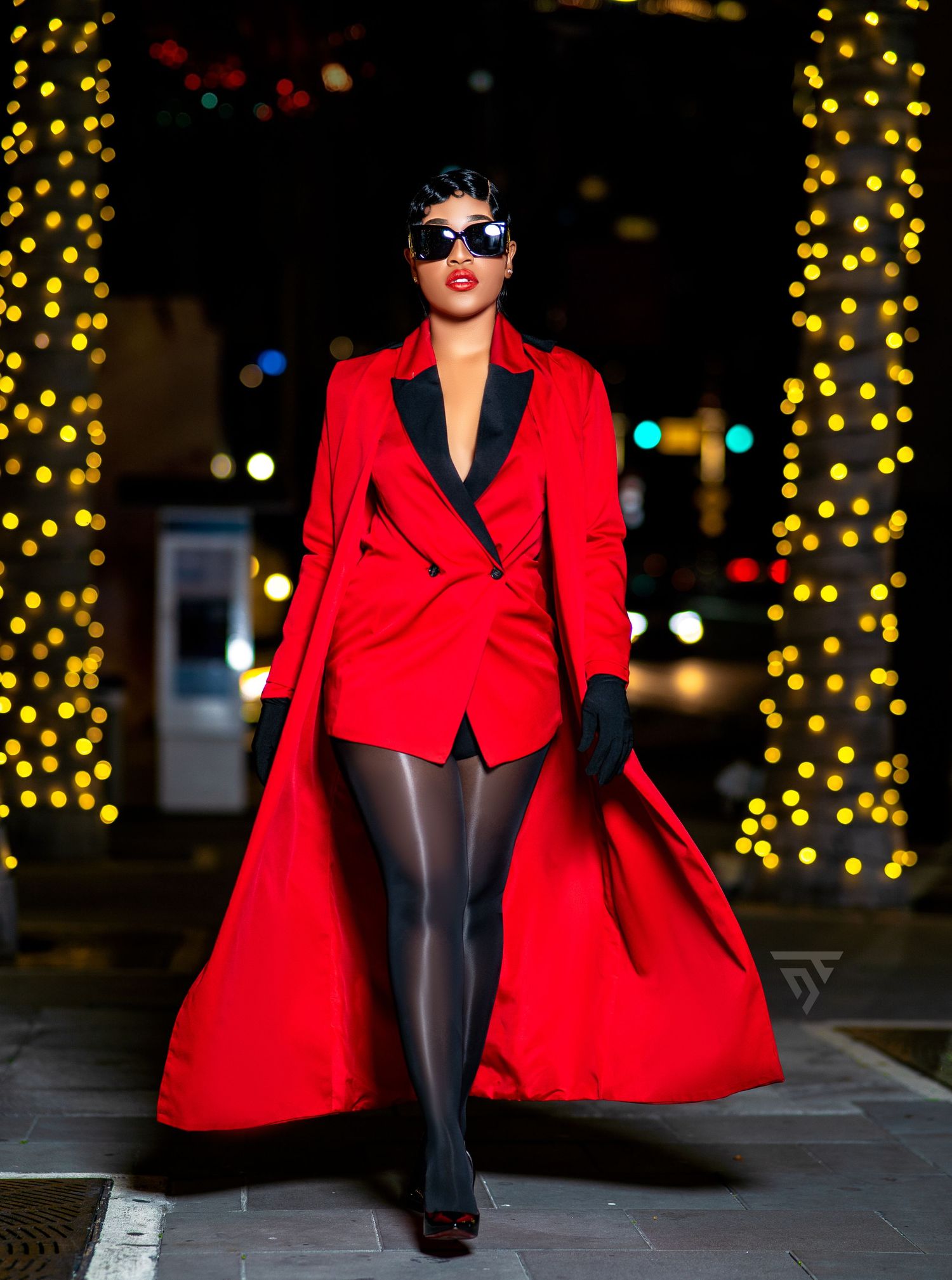 Fashion model in red blazer and black tights poses against backdrop of yellow holiday lights.