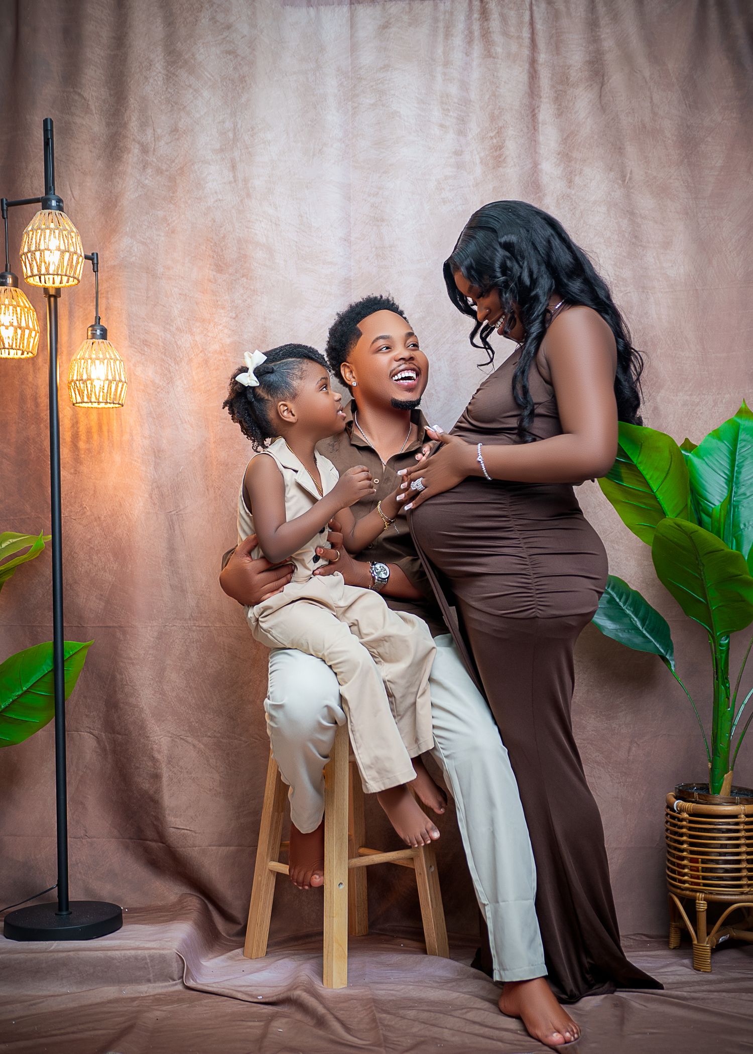 A joyful maternity photo of a pregnant woman in a brown dress with her partner and young daughter in a studio setting.