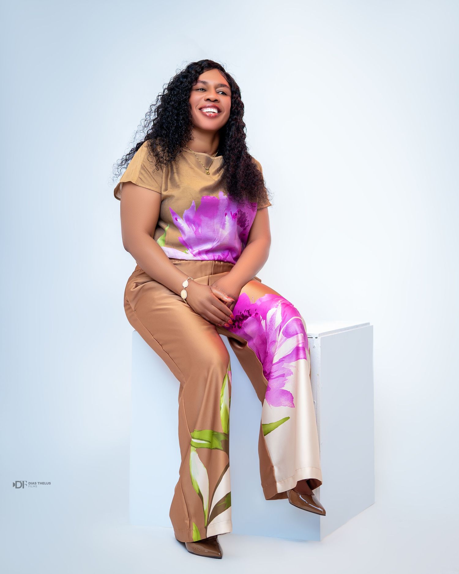 Woman in colorful floral top and tan pants sitting on white cube, smiling confidently in studio photo.