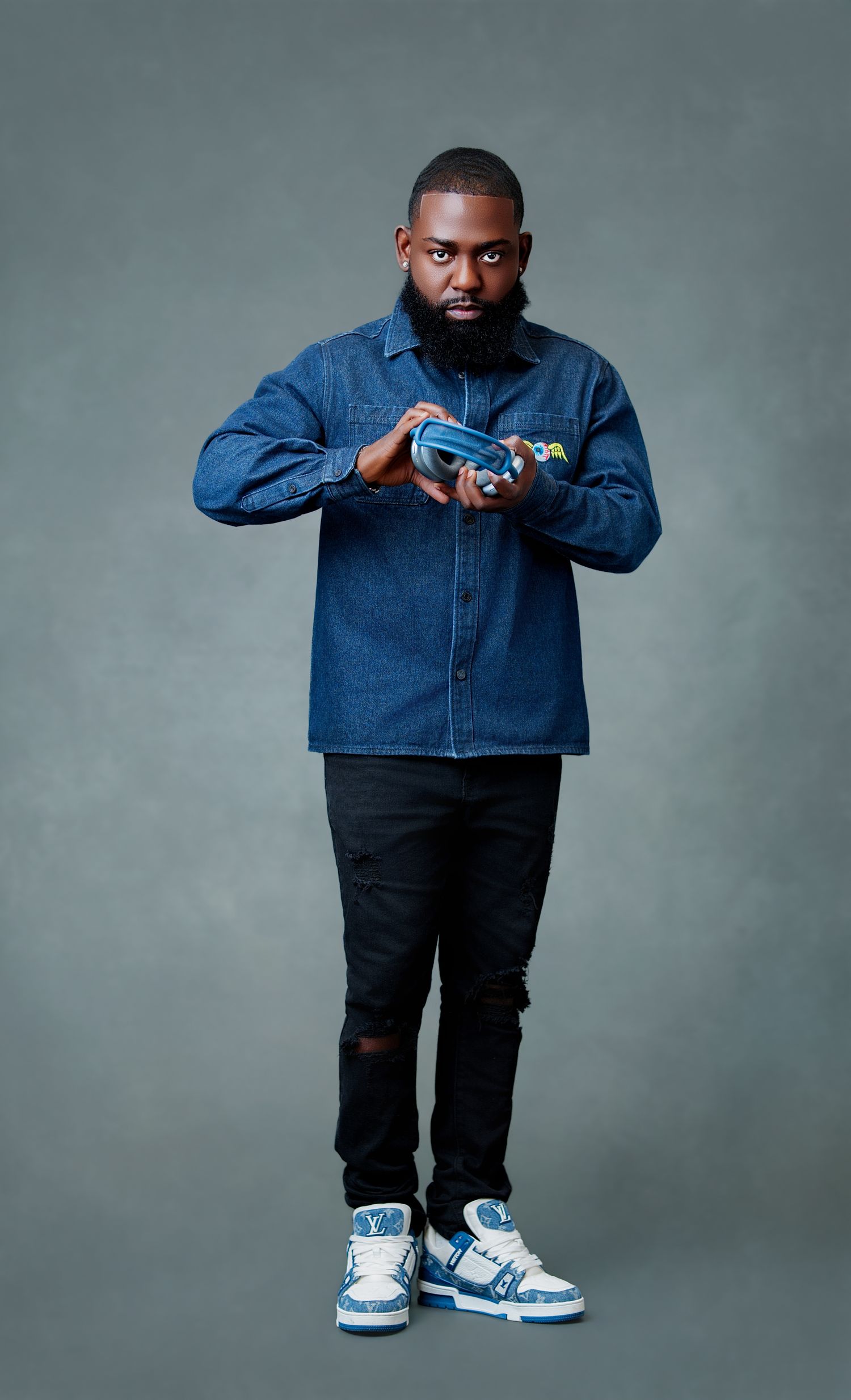 Man in denim jacket, black jeans, and blue sneakers posing confidently against a gray studio background.