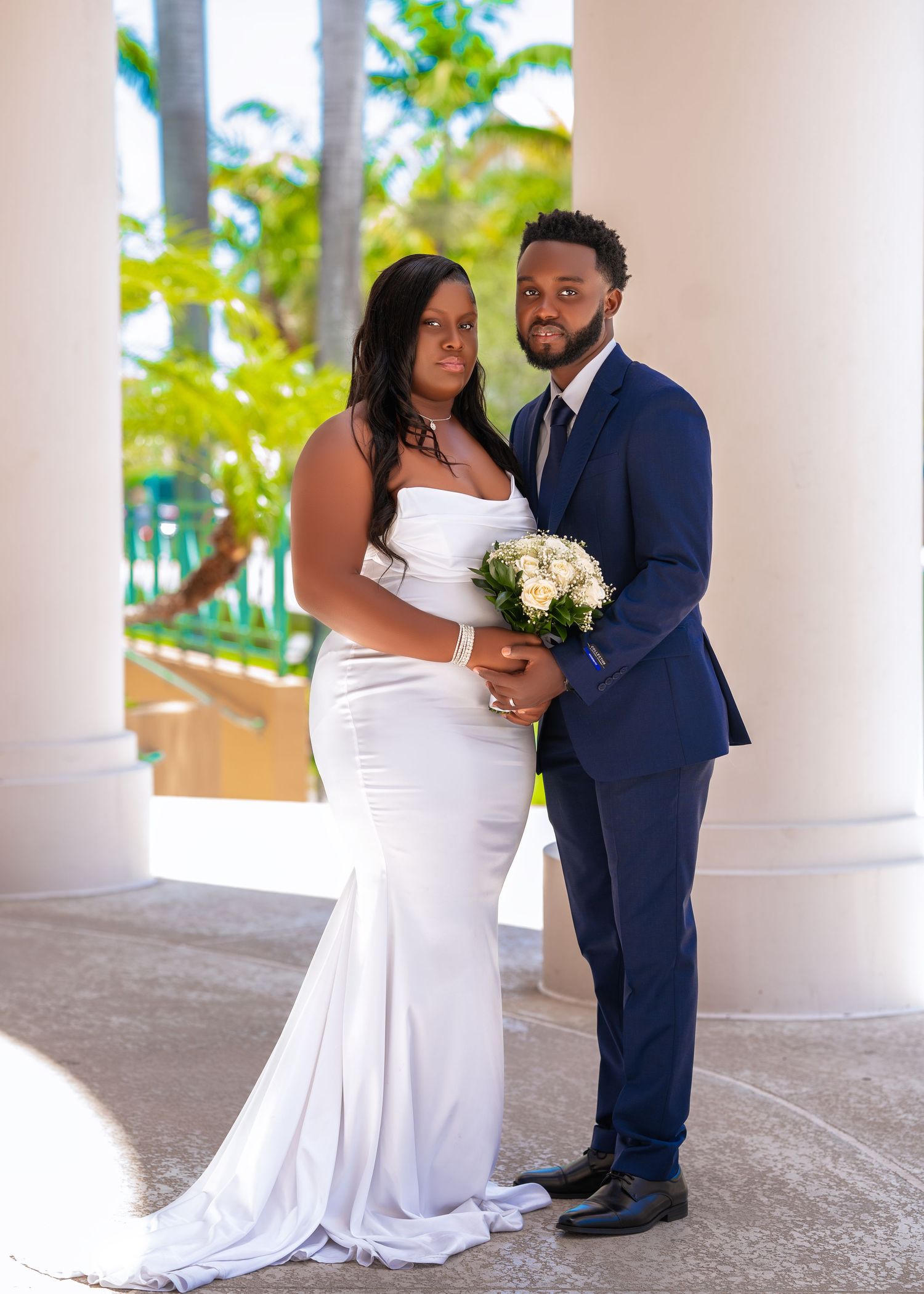 Elegant wedding couple poses together by tropical columns in navy suit and white gown.