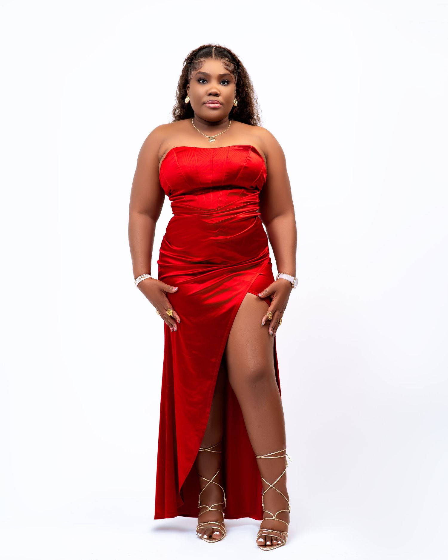 Woman in red strapless satin gown with thigh-high slit and strappy heels posing against white background.