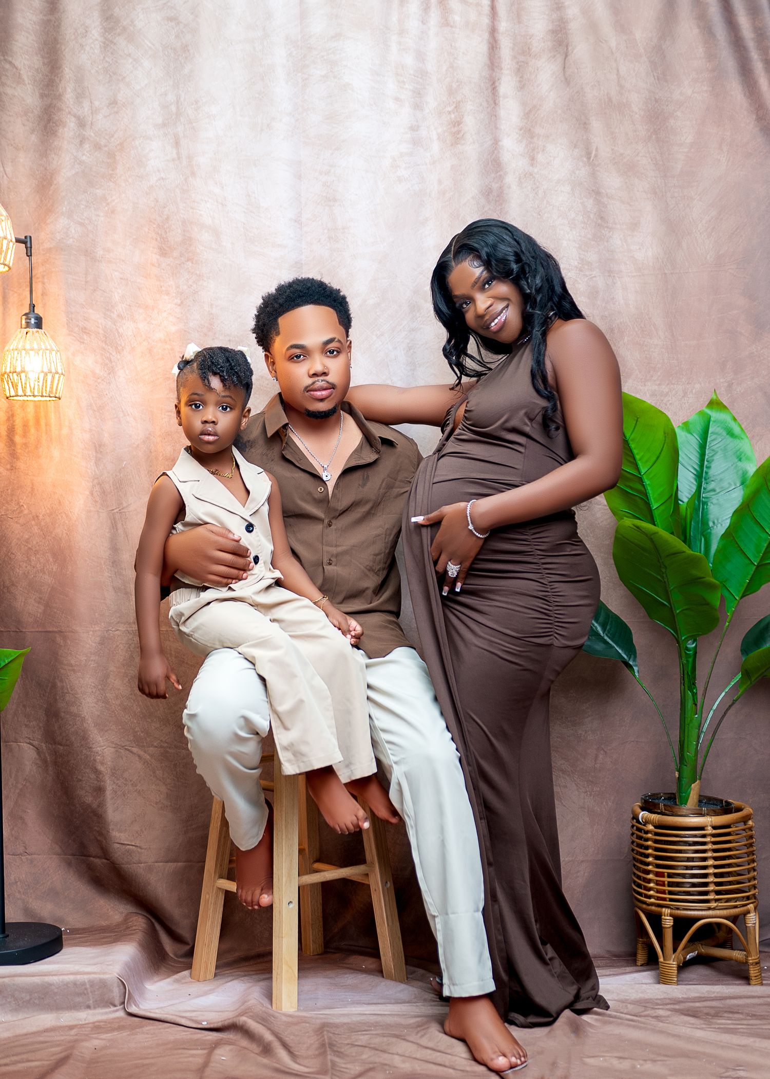 A family maternity photo with a pregnant woman in a brown dress, a man seated on a stool, and a young child posing together.