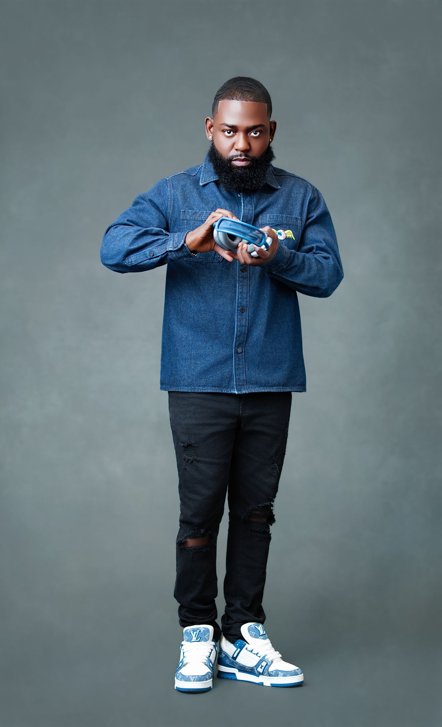 Man in denim jacket, ripped black jeans, and blue high-top sneakers posing confidently against a gray background.