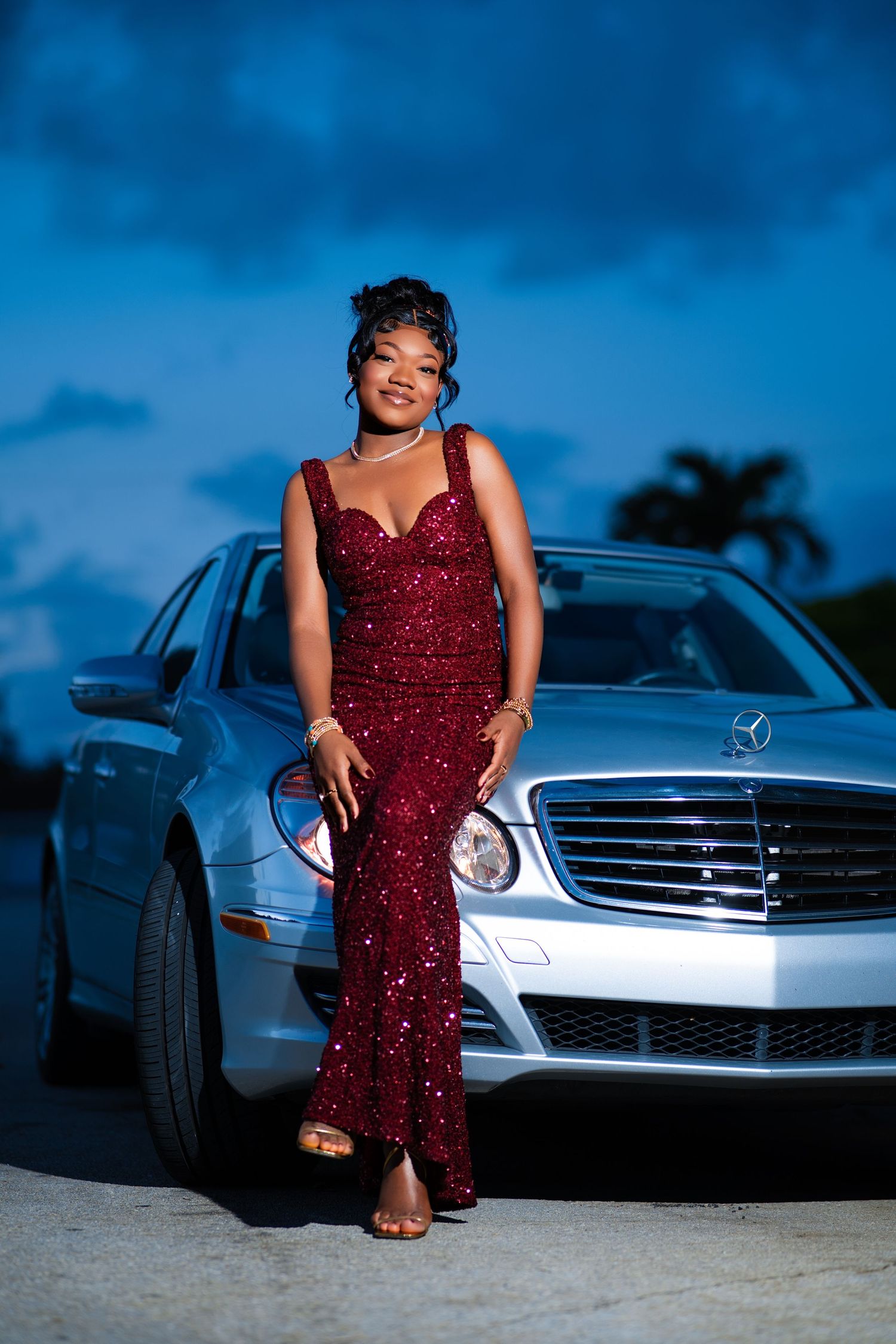 Elegant woman in burgundy sequin gown poses beside silver Mercedes-Benz sedan at twilight.
