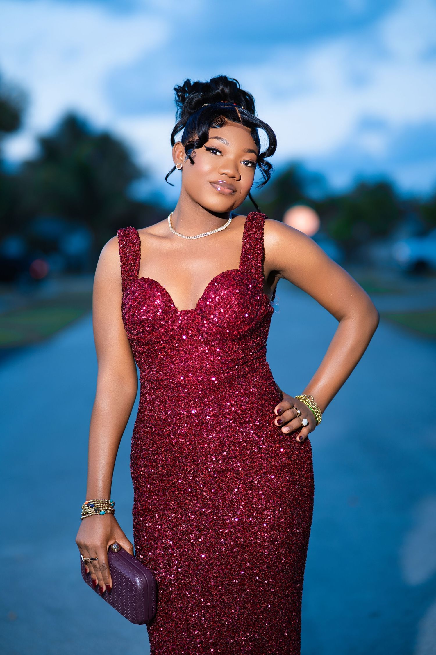 Young woman in a sparkling burgundy sequin gown holding a clutch purse, posing outdoors at dusk.