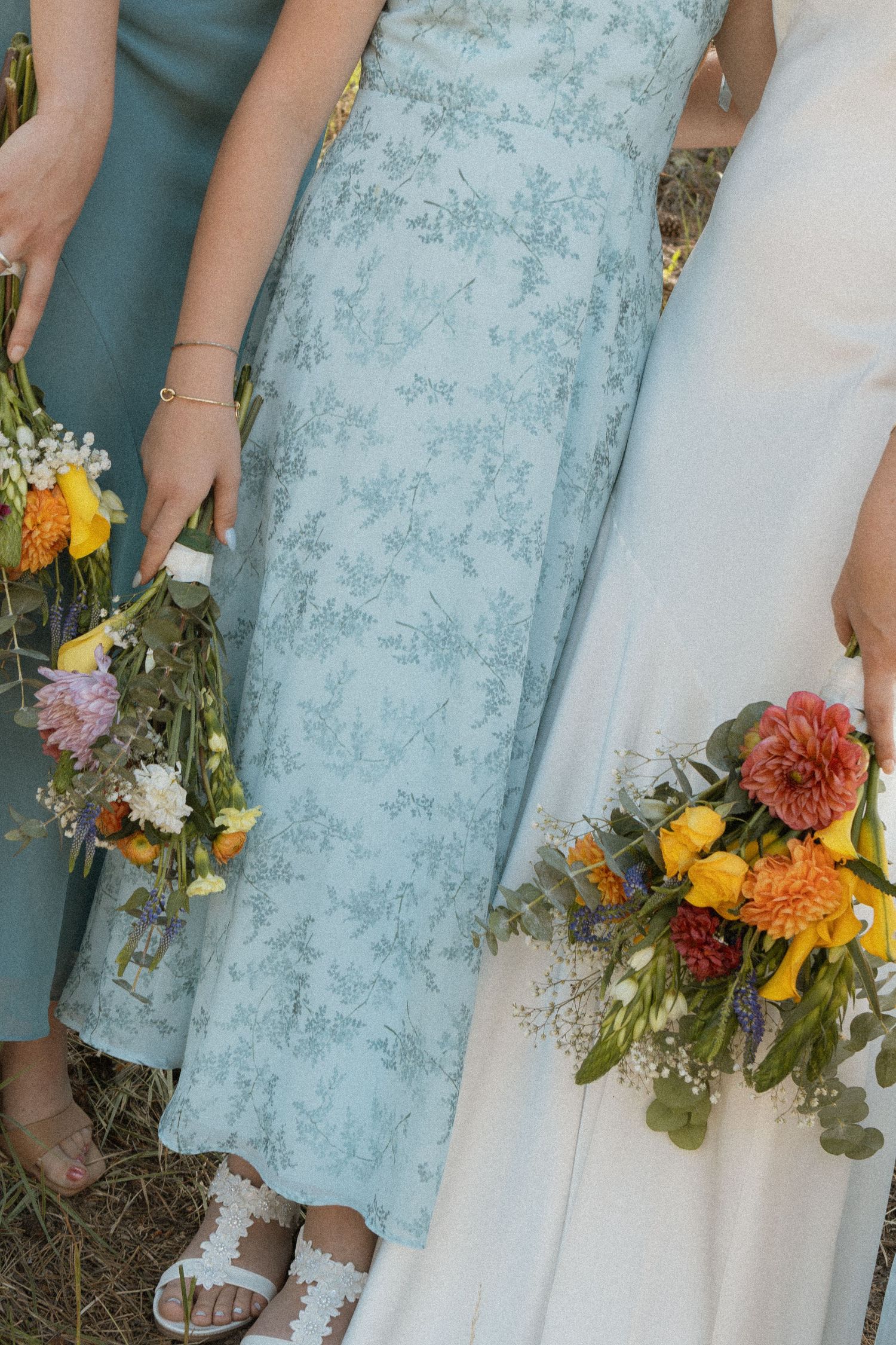Bridesmaids in sage green dresses holding colorful wildflower bouquets beside a bride in white at an outdoor wedding.