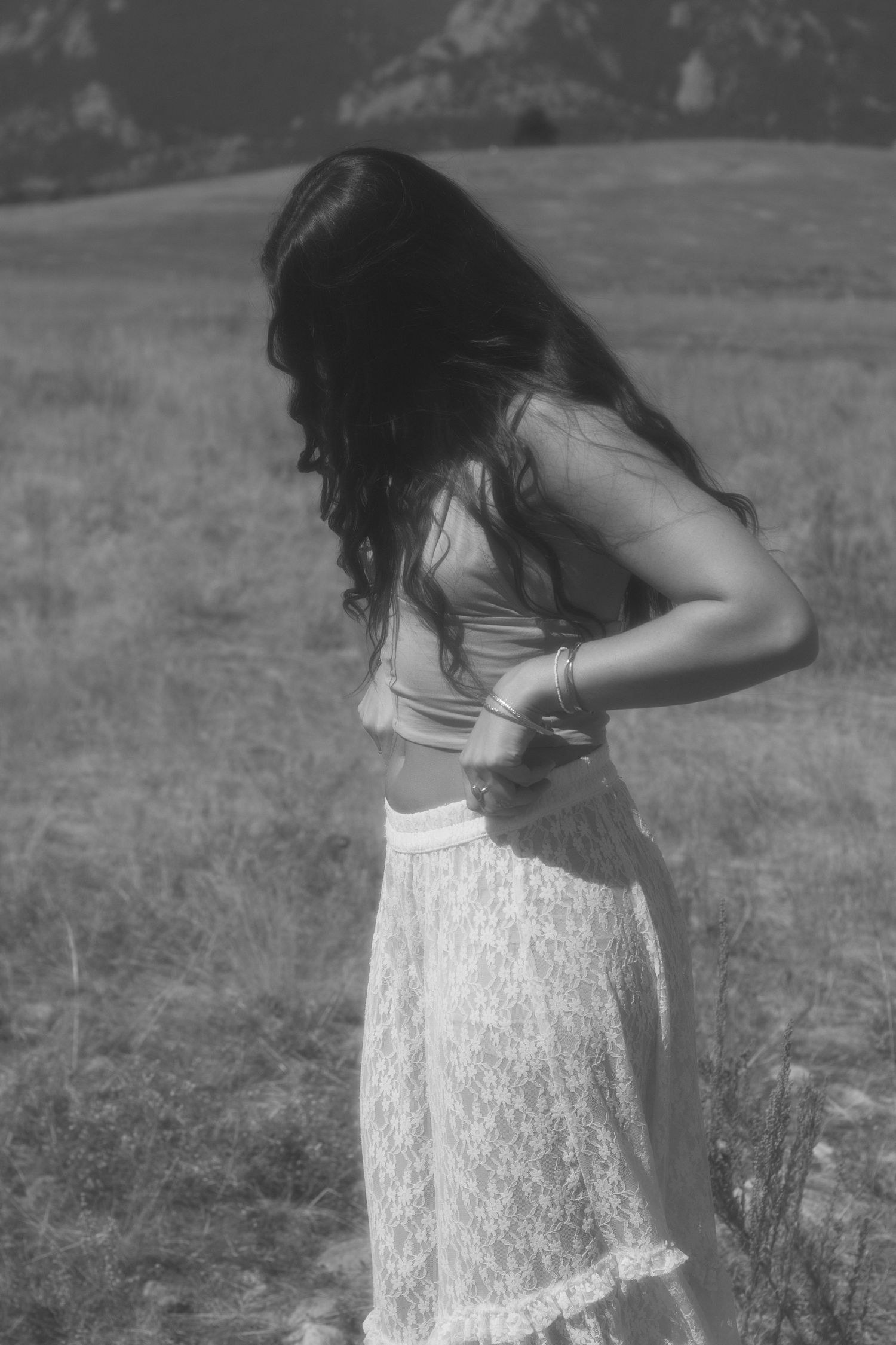 Figure in white lace dress poses elegantly in a mountain field during golden hour.